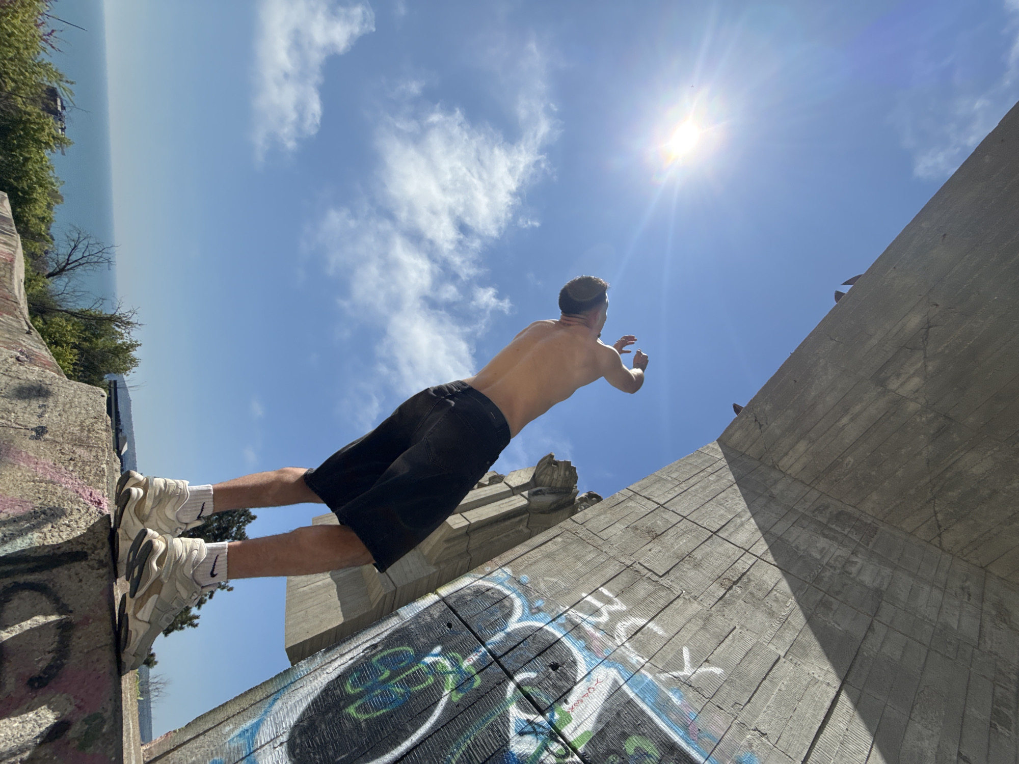 A shirtless man in black shorts and white sneakers is jumping from a concrete structure, likely a bridge or building, with graffiti on its surfaces. The man's feet are on the edge of one section of the structure, and his body is extended horizontally towards the sun and blue sky. The sea and treeline are visible in the background, suggesting a coastal location. Based on the style of the concrete structure and surrounding landscape, the location is likely the Asparuhov Bridge in Varna, Bulgaria. The sun is high in the sky, indicating it is daytime. There are two small birds on the right edge of the structure.