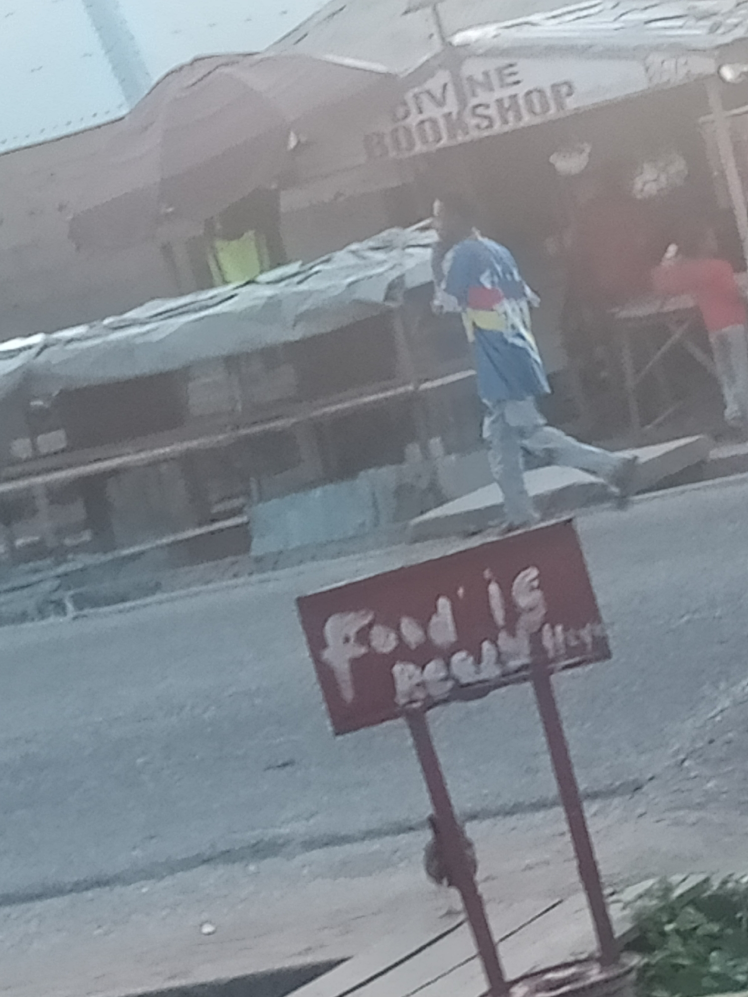 In Jalingo, Nigeria, a person in a colorful shirt and jeans runs past a red sign that reads "Food is Ready." In the background, a building displays the words "Divine Bookshop" and appears to be covered by tarpaulin. The scene is captured on a street with a gray, paved surface.