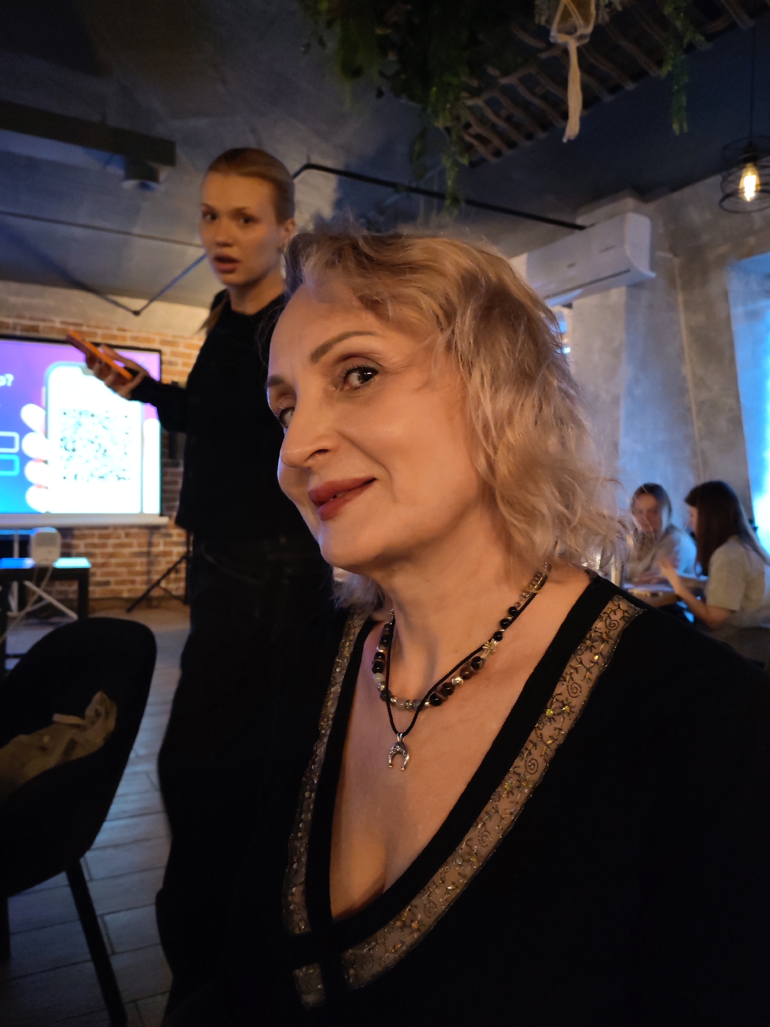 An indoor scene in Chaikovsky, Russia, depicts an adult female in the foreground, primarily facing to her right with a slight smile, positioned in front of a blurred background featuring multiple individuals and presentation equipment.
The foreground subject is a fair-skinned adult female with wavy, light-colored hair, wearing a black V-neck top with an embroidered gold-patterned trim. She wears a multi-strand beaded necklace, one strand featuring dark brown and lighter beads, and another thinner black cord with a silver-colored horseshoe pendant.
Behind her, slightly to the left, a second adult female with light hair pulled back is visible, wearing a dark top. She holds an orange smartphone horizontally in her right hand, gesturing towards a projected image on a white screen. The projection displays a large QR code on a purple and blue gradient background, with partially visible text including a question mark and a phone icon. The second female's mouth is open, suggesting she is speaking or reacting.
In the blurred background to the right, two additional individuals, likely females, are seated at a table. One wears a light-colored top, the other a darker one. This area is illuminated by a subtle blue glow.
The environment features a dark ceiling with visible structural elements and hanging plant decorations. An air conditioning unit is mounted on a light grey wall. A modern light fixture with an exposed bulb is visible on the upper right. A brick wall section is present behind the projection screen. A dark, possibly upholstered chair is partially visible in the lower left. The overall lighting is dim, suggesting an evening or intentionally moody setting.