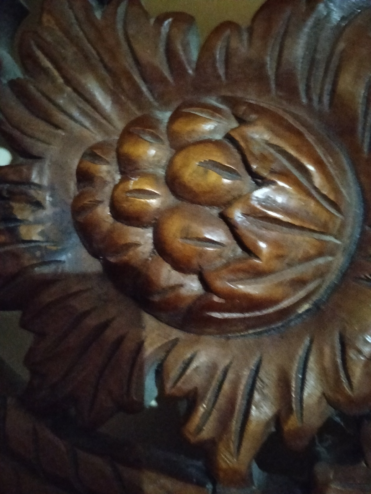 This is a close-up, low-light photograph of carved wooden ornamentation, likely part of furniture or a decorative architectural element. The carving depicts a stylized floral or fan-like motif with rounded, overlapping petal shapes at the center, which are themselves grooved to suggest smaller segments. These central elements are surrounded by radiating, elongated shapes with deep grooves, resembling petals or leaves. The wood is a dark brown with a polished finish that reflects light, highlighting the carved contours and creating a sense of depth. The overall texture is detailed and artisanal. The image is cropped tightly, focusing on the texture and form of the carving, with no discernible background elements that would indicate a specific location within Payagaboh, Indonesia.