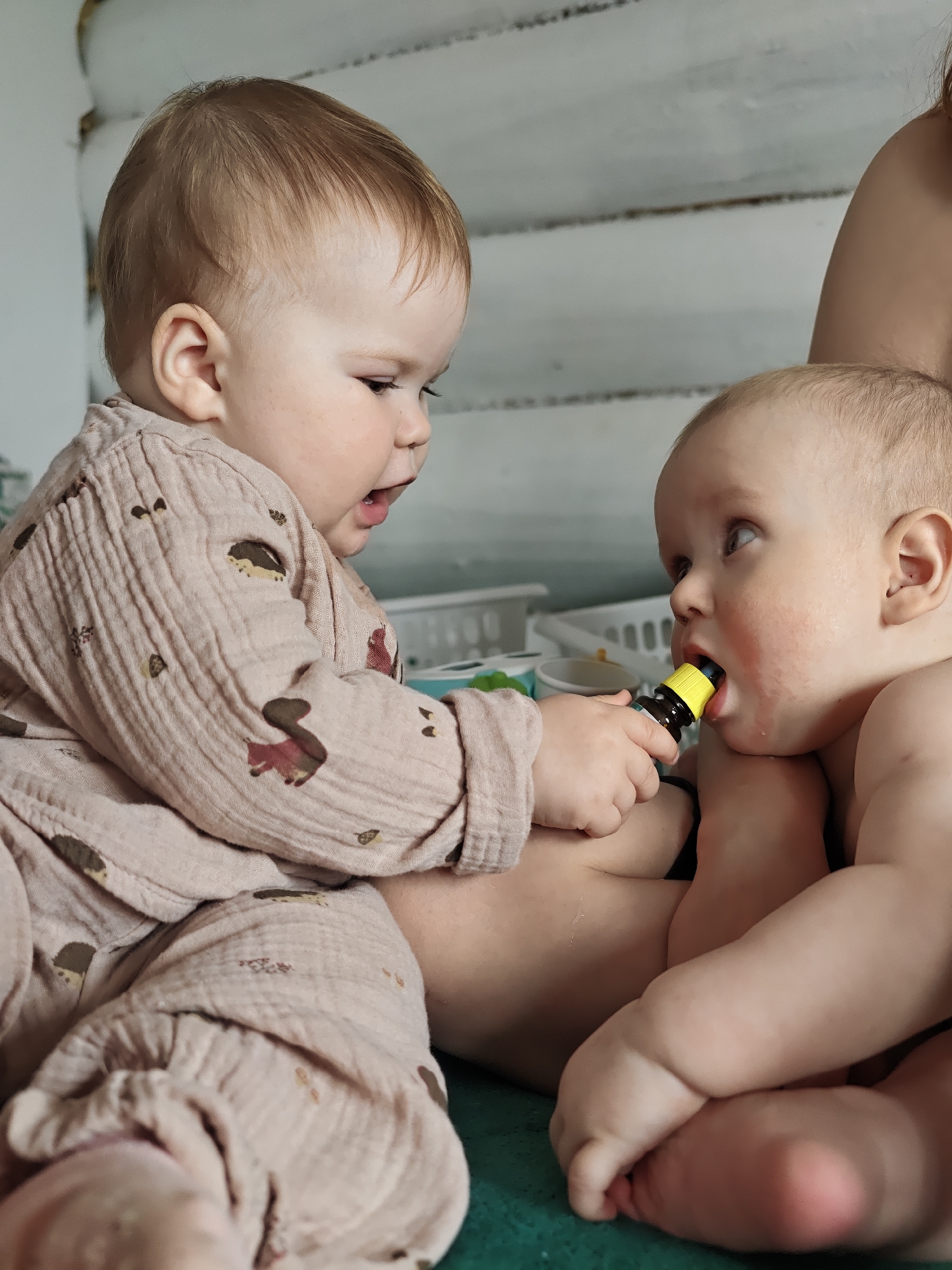 Two babies are captured in a close-up, indoor shot, engaged in an interaction. The baby on the left wears a light brown or pink long-sleeved romper made of crinkled fabric, adorned with small brown hedgehog and squirrel patterns. This baby has light brown hair and an open mouth, appearing to be speaking or expressing surprise, looking intently at the other baby.
The baby on the right is bare-chested, with fair skin and light blonde hair. This baby holds a small, dark brown bottle with a bright yellow cap or nipple in its mouth, looking upwards and to the left. The left baby's hand appears to be guiding or presenting the bottle to the right baby. An adult's shoulder or arm is partially visible behind the right baby, suggesting they are being held or supported.
The background reveals a light-colored, possibly white, wooden plank wall and hints of white plastic containers or baskets, indicating an indoor domestic setting. The babies are positioned on a green surface, likely a mat or blanket. The overall mood is one of curious engagement and playful interaction between the two infants. There is no visible text in the image. This scene takes place in Noviy, Russia.