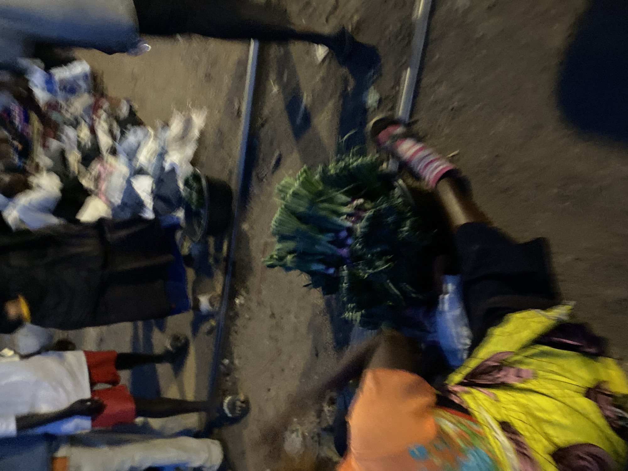 The image appears to show a scene in Bukuru, Nigeria. Several individuals are present, some lying down or sleeping on what seems to be a street or ground surface. Some individuals are wearing casual clothing. Bundles of green vegetables are seen near one person. Objects such as a cart or trolley-like structure are observed, holding items appearing to be merchandise or goods. Other objects include clothing items. The environment seems to be a street setting, likely at night or in low-light conditions.