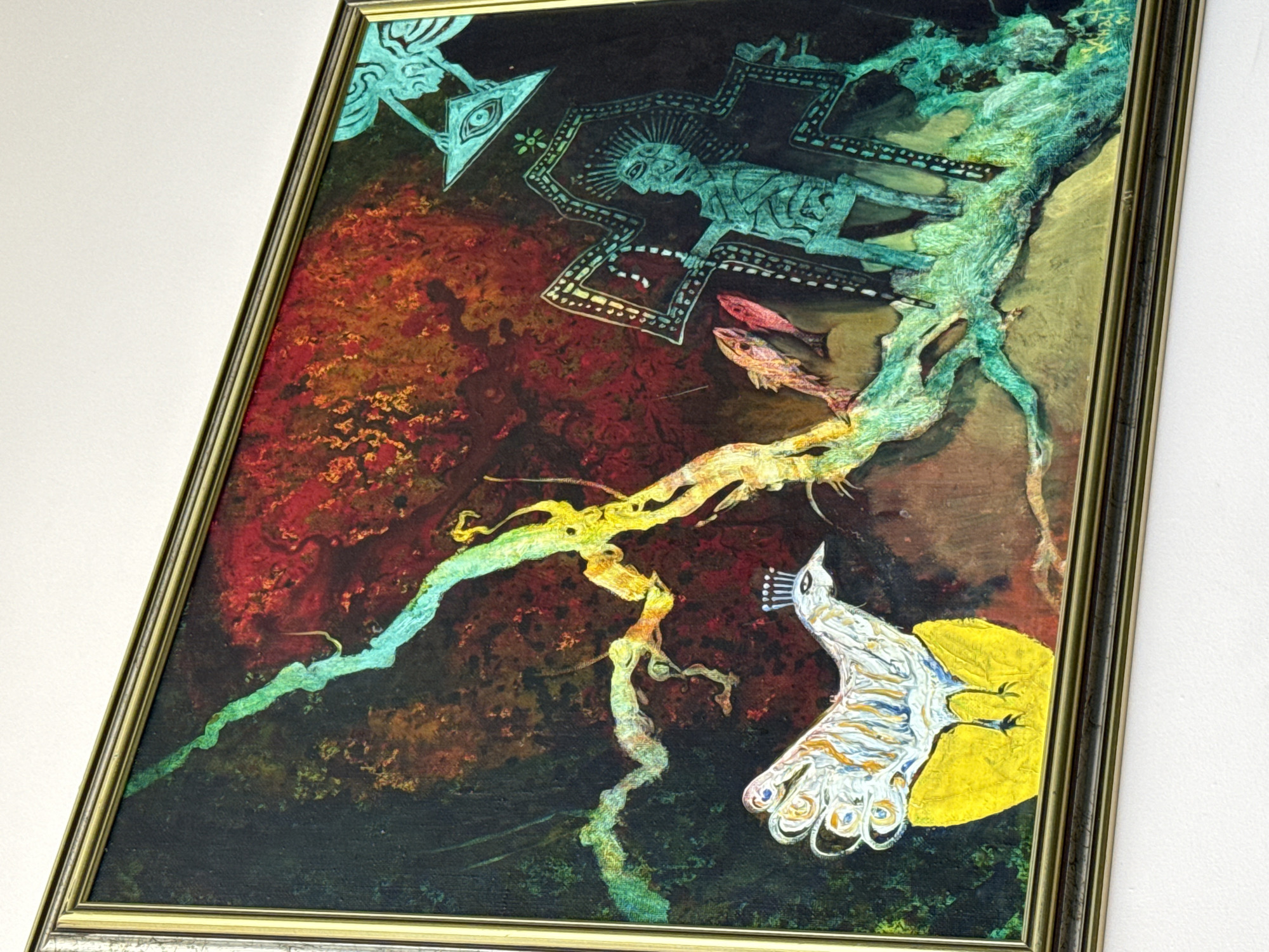The image shows a framed painting hanging on a white wall. The painting features a surreal scene with a dark background transitioning from reddish hues at the top to a dark black at the bottom. In the upper part of the painting, there are stylized figures reminiscent of indigenous art or hieroglyphs, depicted in a light blue or teal color. Two fish swim between these figures. An abstract, branching structure with a range of colors (green, yellow, orange) extends diagonally downwards across the canvas. In the lower right corner, there is a white bird with a yellow circle behind it. No specific location in Plovdiv, Bulgaria, is evident in the image.