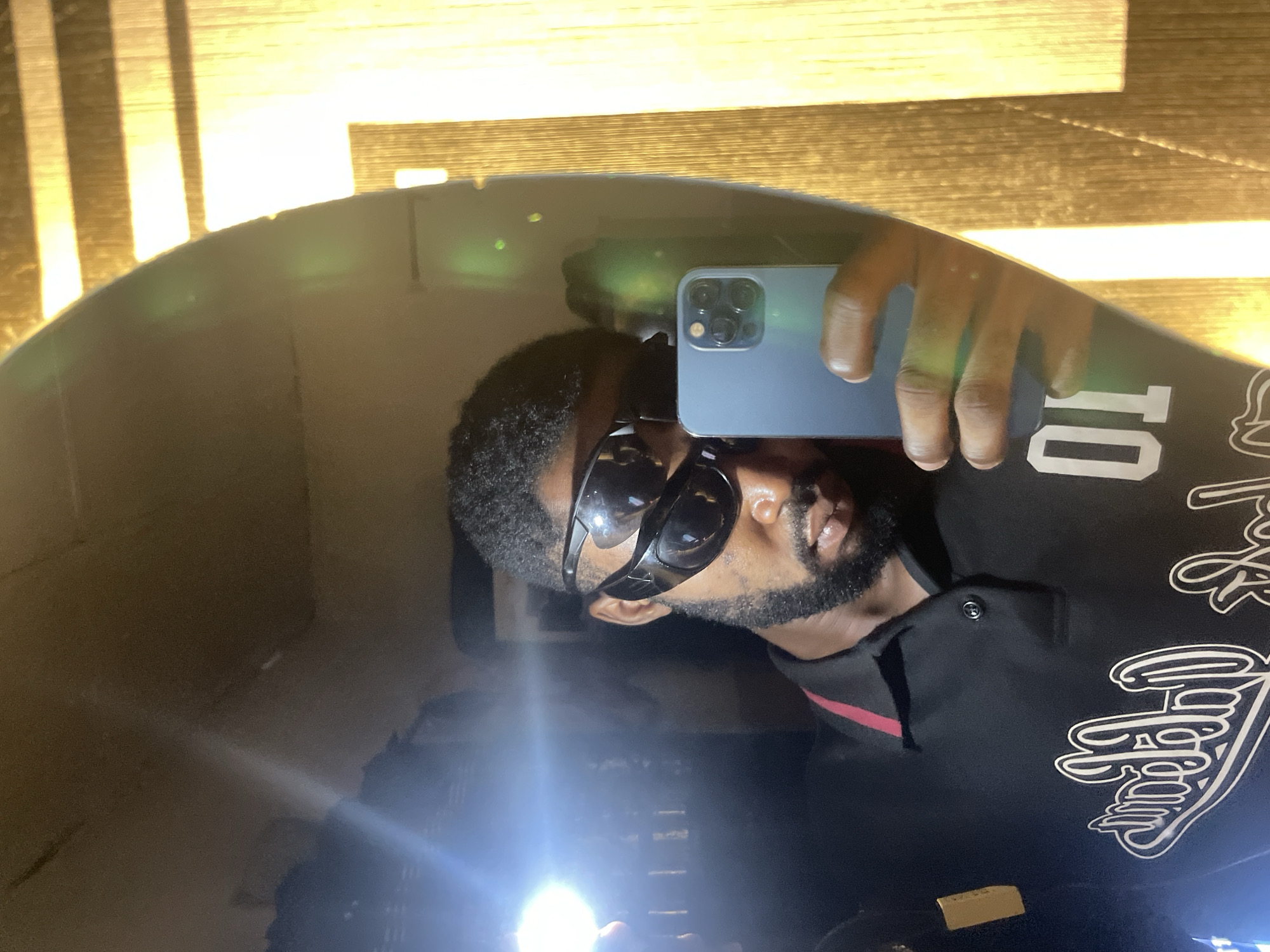 A dark-skinned male adult with a beard and short dark hair is depicted taking a mirror selfie in an indoor setting in Lokoja, Nigeria. He wears two pairs of black wraparound sunglasses, one stacked over the other, obscuring his eyes. He is dressed in a black polo shirt with a red trim on the collar. The shirt features reversed white stylized text and the reversed number "01" on the chest. The man holds a light blue/grey smartphone with a visible triple-lens camera module in his right hand, angled to capture his reflection. A very bright, circular light source, originating from the left side of the frame, casts a strong glare and illuminates the subject. The reflected background shows a light brown wall and a golden-brown vertical paneling or furniture piece with horizontal stripes on the right side.
