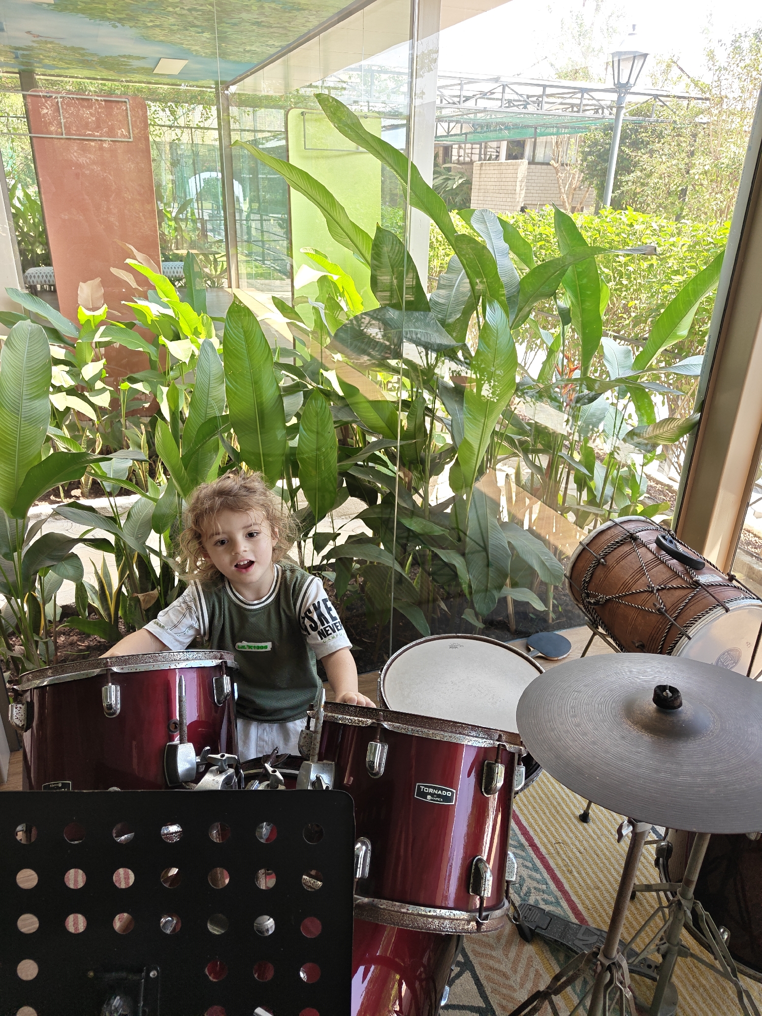 A child with curly hair stands behind a burgundy drum set inside a glass-walled room filled with large, leafy green plants. The drums, labeled "Tornado by Mapex", are partially visible. The room is in Kalote Mokashi, India, as deduced from the metadata. The backdrop visible through the glass includes a building and greenery.