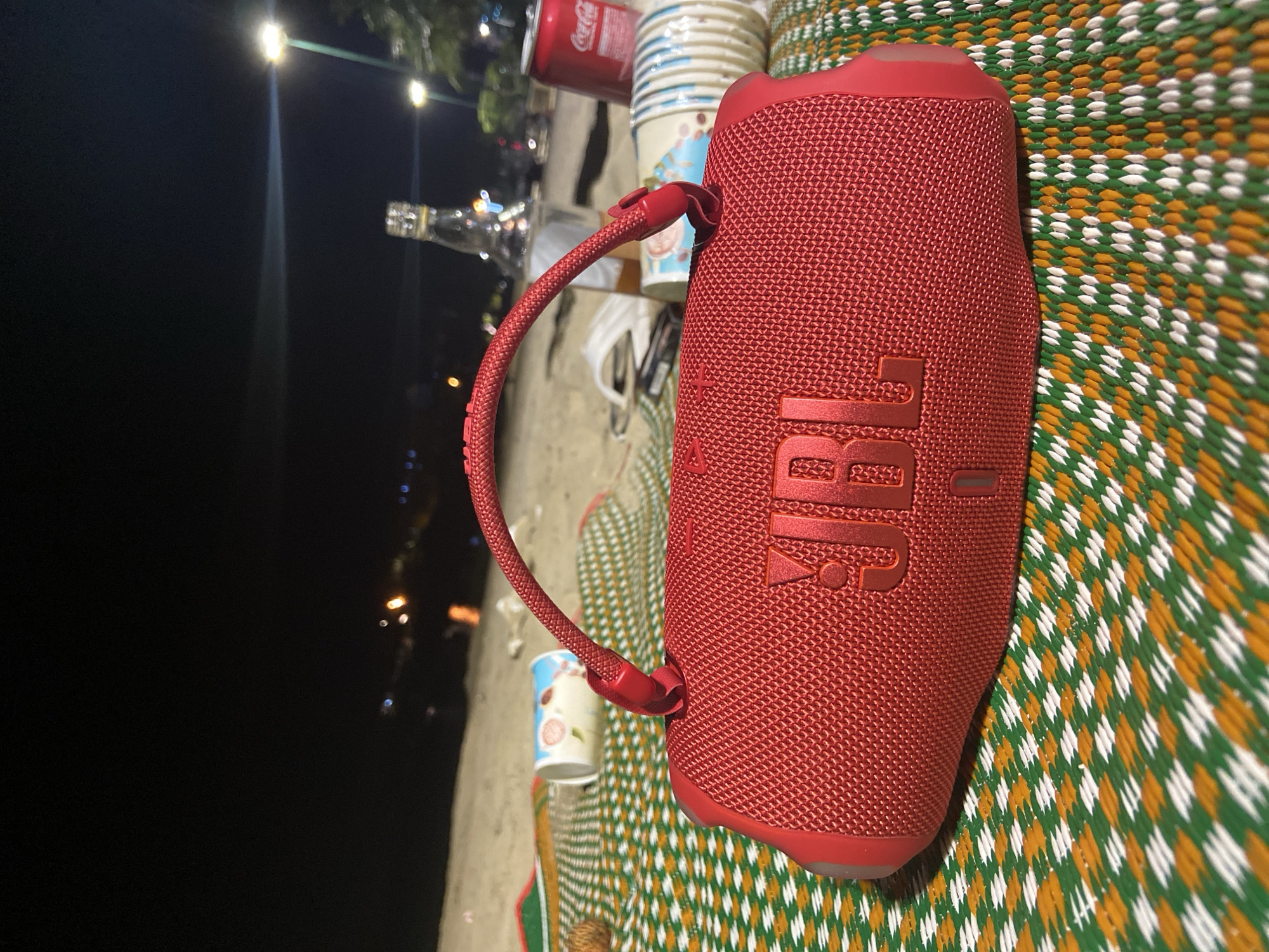A red JBL speaker sits on a woven mat on a beach at night in the city of Nha Trang, Vietnam. In the background, a bottle and a can of Coca-Cola are visible, along with several paper cups. Lights from the city and streetlights illuminate the scene. The water of the sea is visible beyond the beach.