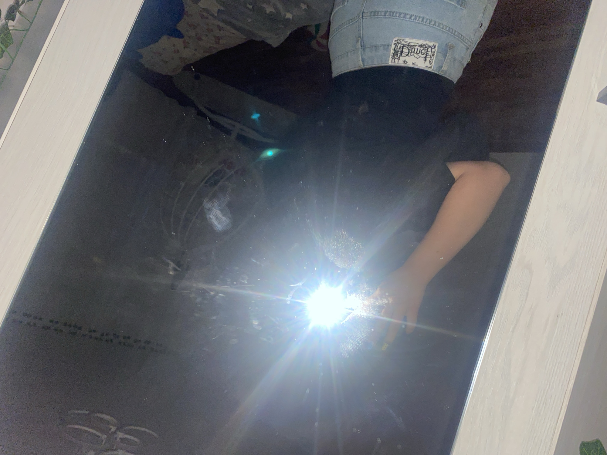 The image displays a mirror selfie taken indoors, with a bright camera flash as the central light source, obscuring direct visual details. The reflected environment offers no discernible features to confirm a location in the city of Noviy, Russia. A person is visible in reflection, holding a smartphone in their right hand, which emits a strong, star-shaped flash. This flash creates significant lens flare and overexposure, making the person's face and upper torso indistinct. The visible portions of the person include a right forearm and hand, wearing a dark ring on one finger. Their left forearm is partially visible to the left of the flash. The person is dressed in a black short-sleeved top and light-wash denim bottoms, possibly jeans or shorts, with a distinct rectangular graphic patch on the back right pocket. The mirror itself is framed by a light-colored, possibly white or light grey, wooden border with visible grain, spanning the top and bottom of the image. The mirror surface exhibits numerous small dust particles and smudges. Reflected in the top-left area of the mirror are faint, abstract circular shapes and a vertical line of small, dark dots. In the darker areas to the right of the central flash, multiple green lens flares are present. Partially visible in the reflection to the right of the person is a patterned fabric, potentially a blanket or cushion, featuring star motifs against a light background. The overall reflected background appears dark and indistinct, suggesting a dimly lit room or dark furnishings.
