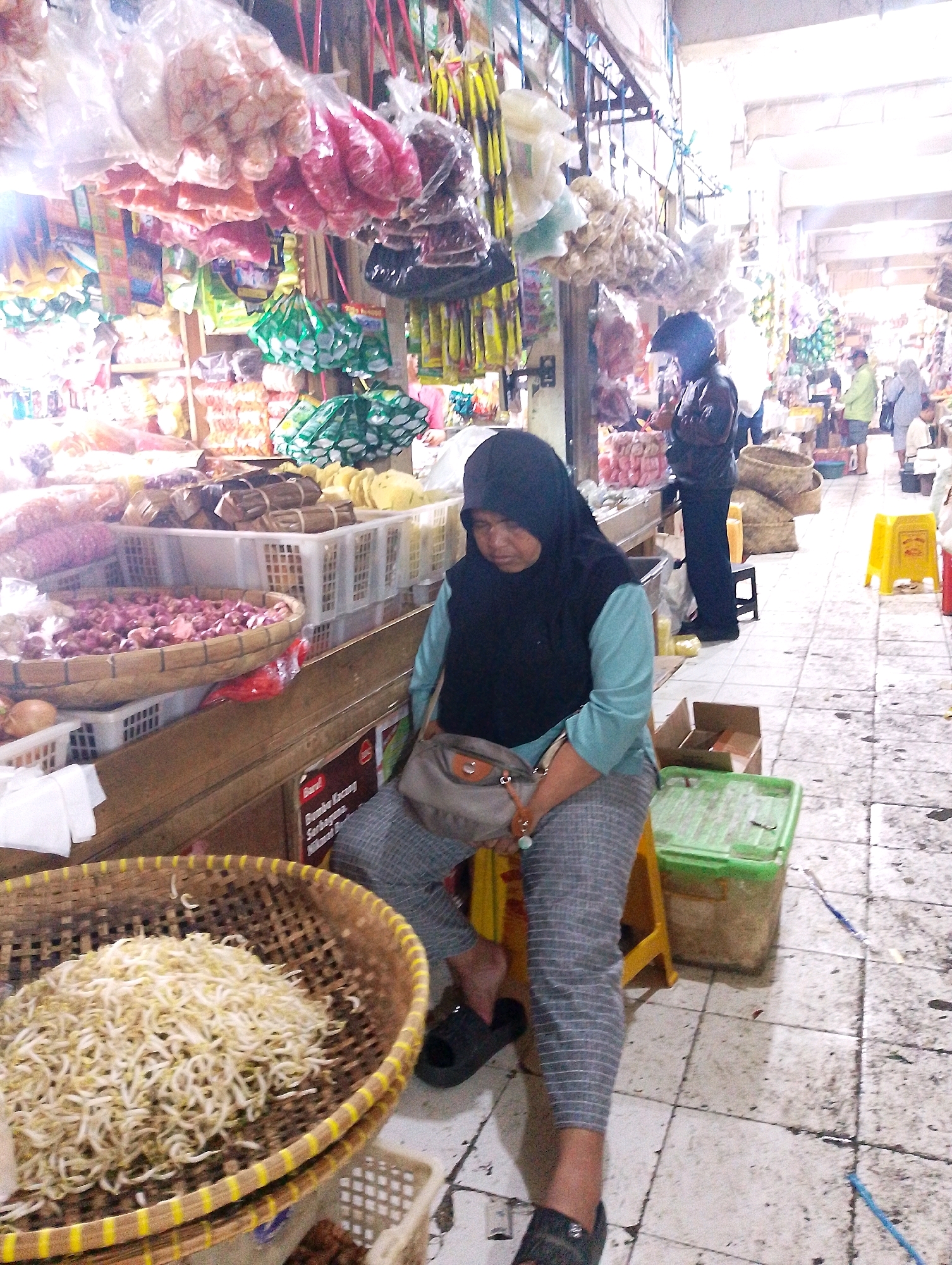A woman wearing a black hijab and a light blue top is sitting on a yellow stool in a market in Lembang, Indonesia. She is wearing grey striped pants and a grey shoulder bag. In front of her, on a wooden counter, is a woven basket filled with bean sprouts. Behind her, hanging from the ceiling, are various goods packaged in plastic bags. The market floor is tiled and appears to be made of stone. Several other people are visible in the background, along with stalls displaying food and other merchandise. A green plastic storage container is on the floor next to the stool.