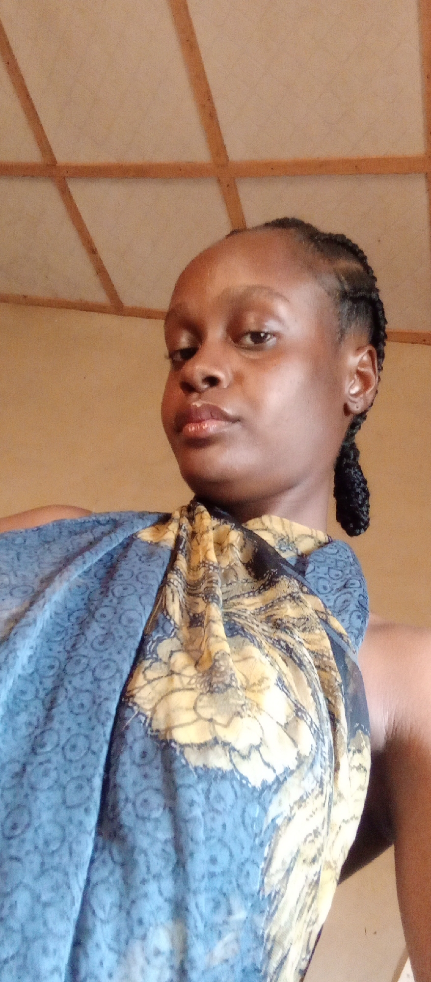 A low-angle medium shot of a Black woman with braided hair and brown skin is wearing a blue and yellow patterned scarf around her shoulders. She is looking at the camera with a serious expression. The background consists of a light brown wall and a ceiling with a wooden grid pattern. Based on the prompt, this image was taken in Akwanga, Nigeria.