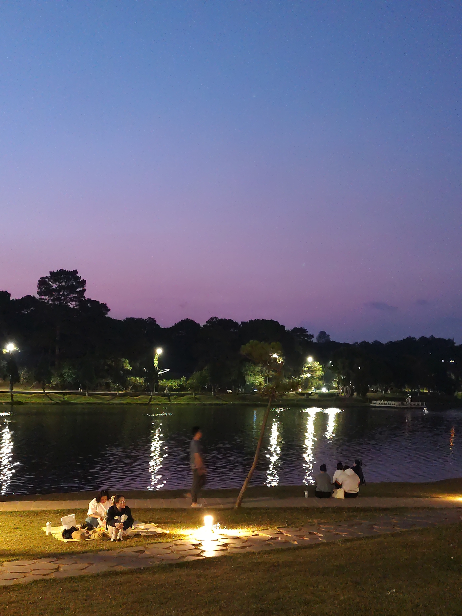 At dusk in Da Lat, Vietnam, a group of people are scattered along the edge of a lake, likely Xuan Huong Lake. In the foreground to the left, two women are sitting on a blanket on the grass, possibly picnicking. Nearby, a single individual is walking. To the right, a small group of people are seated near the water's edge. The lake reflects the lights from the park. Trees line the far shore. The sky is a gradient of purple and blue.
