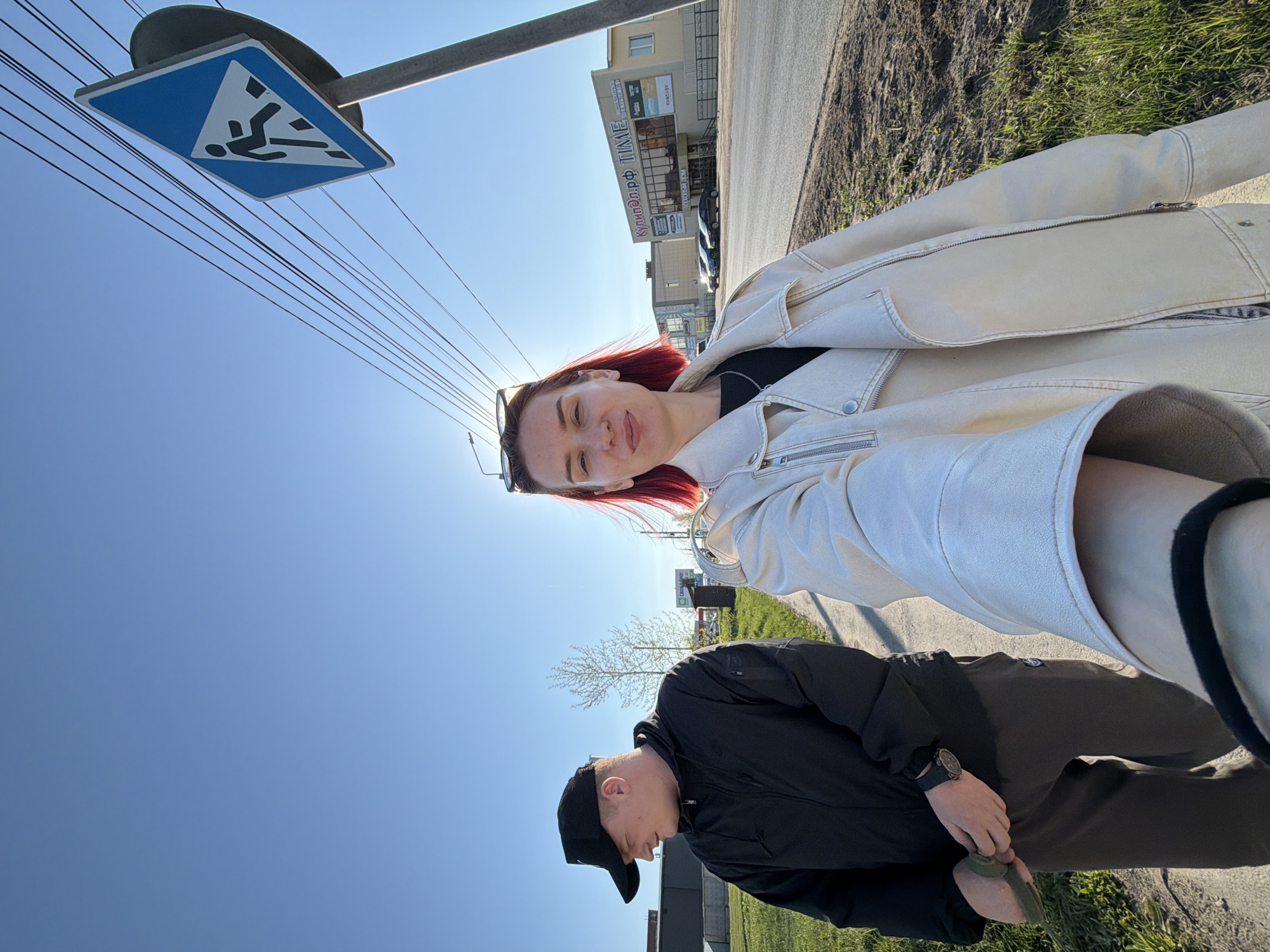 On a sunny day in Chaikovsky, Russia, two individuals are visible on a sidewalk next to a road. The image appears to be a selfie taken at an angle. A woman with red hair and sunglasses is wearing a white jacket. A man in a black jacket and cap is walking behind her. A pedestrian crossing sign is visible in the upper left. Across the road is a building with signage in Russian, one which includes the word "TIME." Grass and a building can also be seen on the side of the road where the pair are standing.