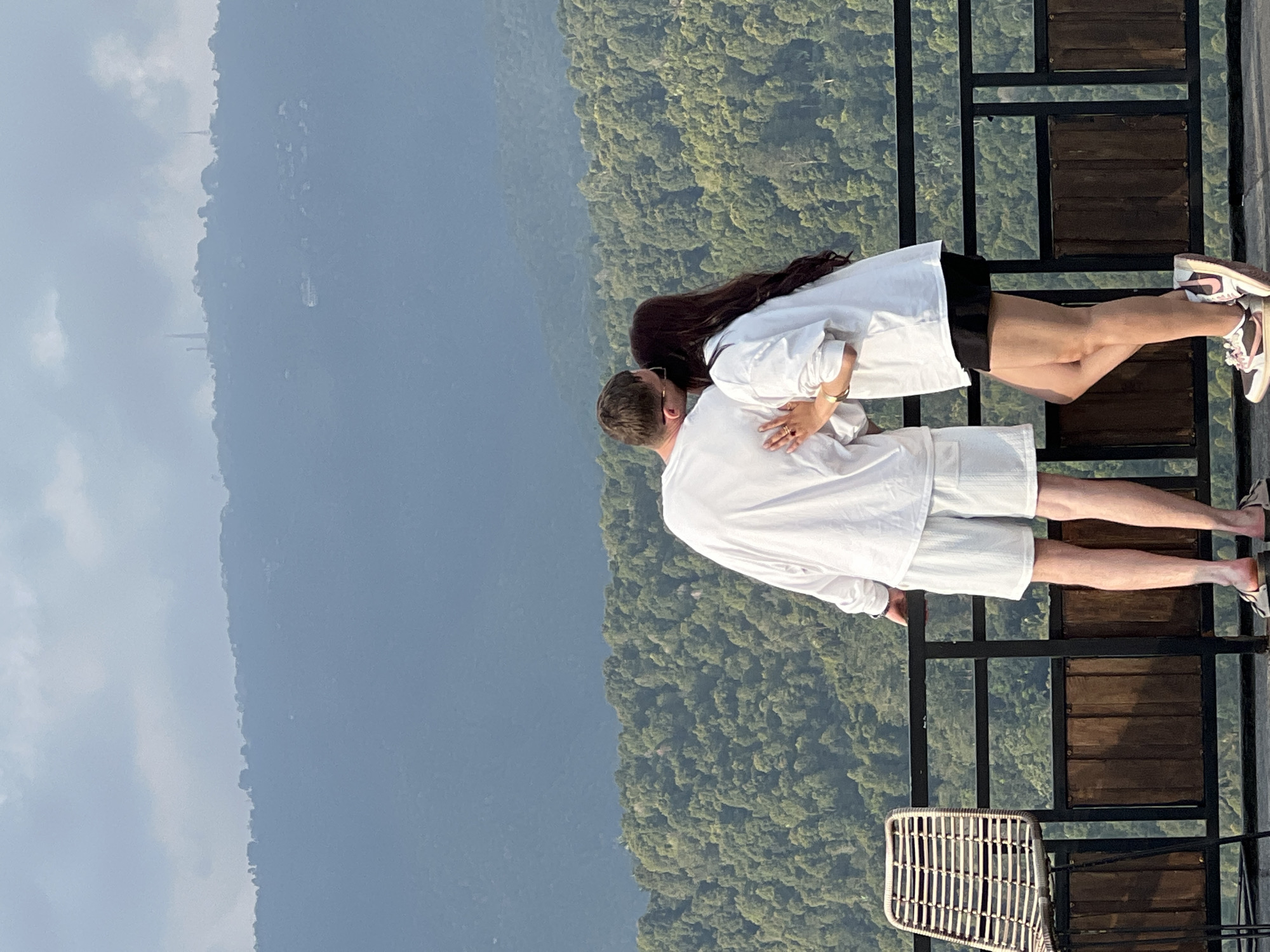 A man and a woman stand on a balcony overlooking a densely forested mountainside in Busungbiu, Indonesia. The man, facing away from the camera, wears a white long-sleeved shirt and white shorts. The woman, also facing away, wears a white long-sleeved shirt and a black skirt. She has her right arm around the man's waist and is leaning in as if to kiss his cheek. The balcony has a black metal railing and a wooden section below. A wicker chair is visible to the left of the frame. The background features rolling green hills and a hazy sky with distant structures visible on the mountain peaks.