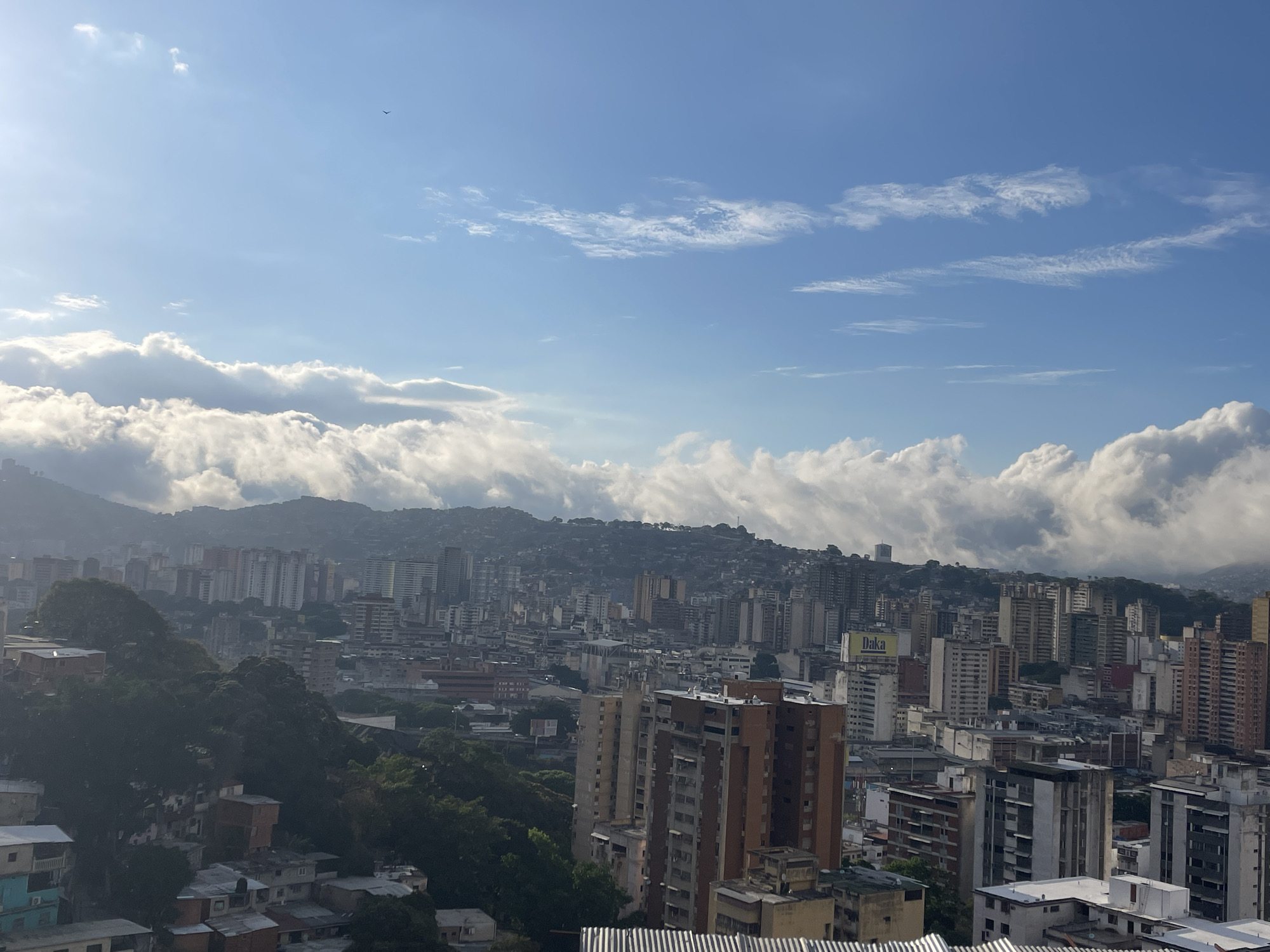 The image depicts a broad daytime cityscape of Caracas, Venezuela, under a sky of bright blue mottled with white clouds. A range of buildings, mostly high-rise apartments, stretch across the view. In the foreground are densely packed, lower-rise residential structures. Behind these, the cityscape rises toward the hills, with taller buildings filling the spaces between the mountain range. The mountains are partially obscured by a thick blanket of low-lying clouds. A single bird is visible in the sky.