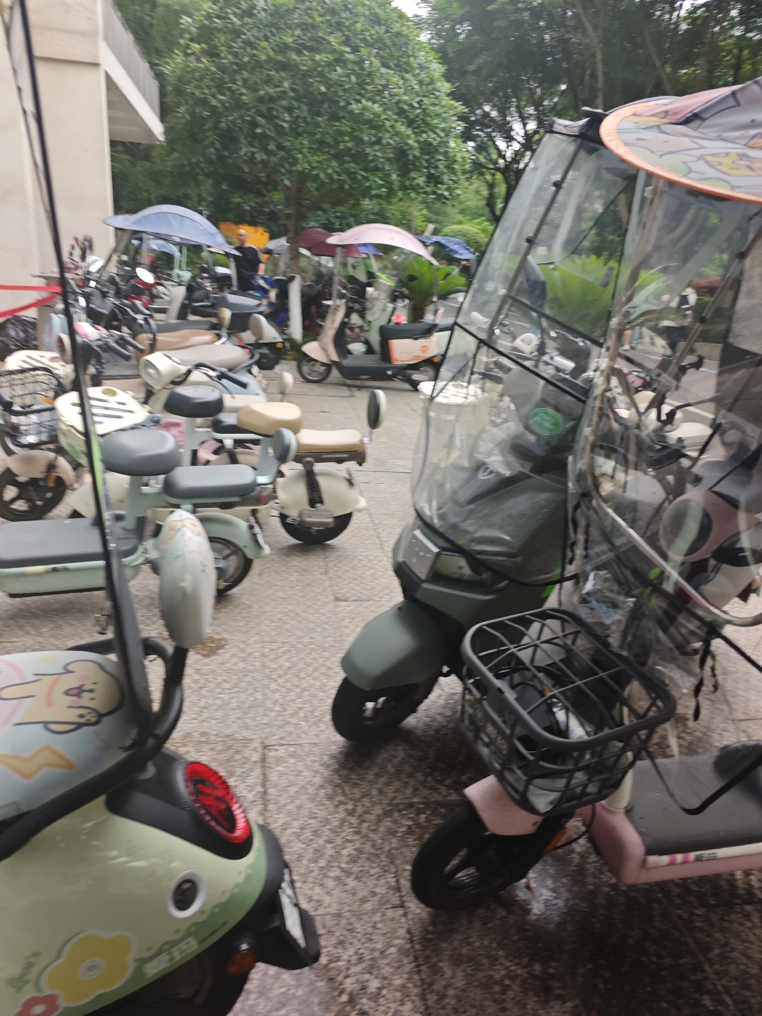 A group of parked electric scooters and motorcycles are visible on a paved surface, likely a sidewalk or parking area, under an overcast sky. Some of the vehicles have transparent covers, suggesting protection from rain. In the background, trees and a building are present. The scene is located in the Chongqing High-tech Zone, China.