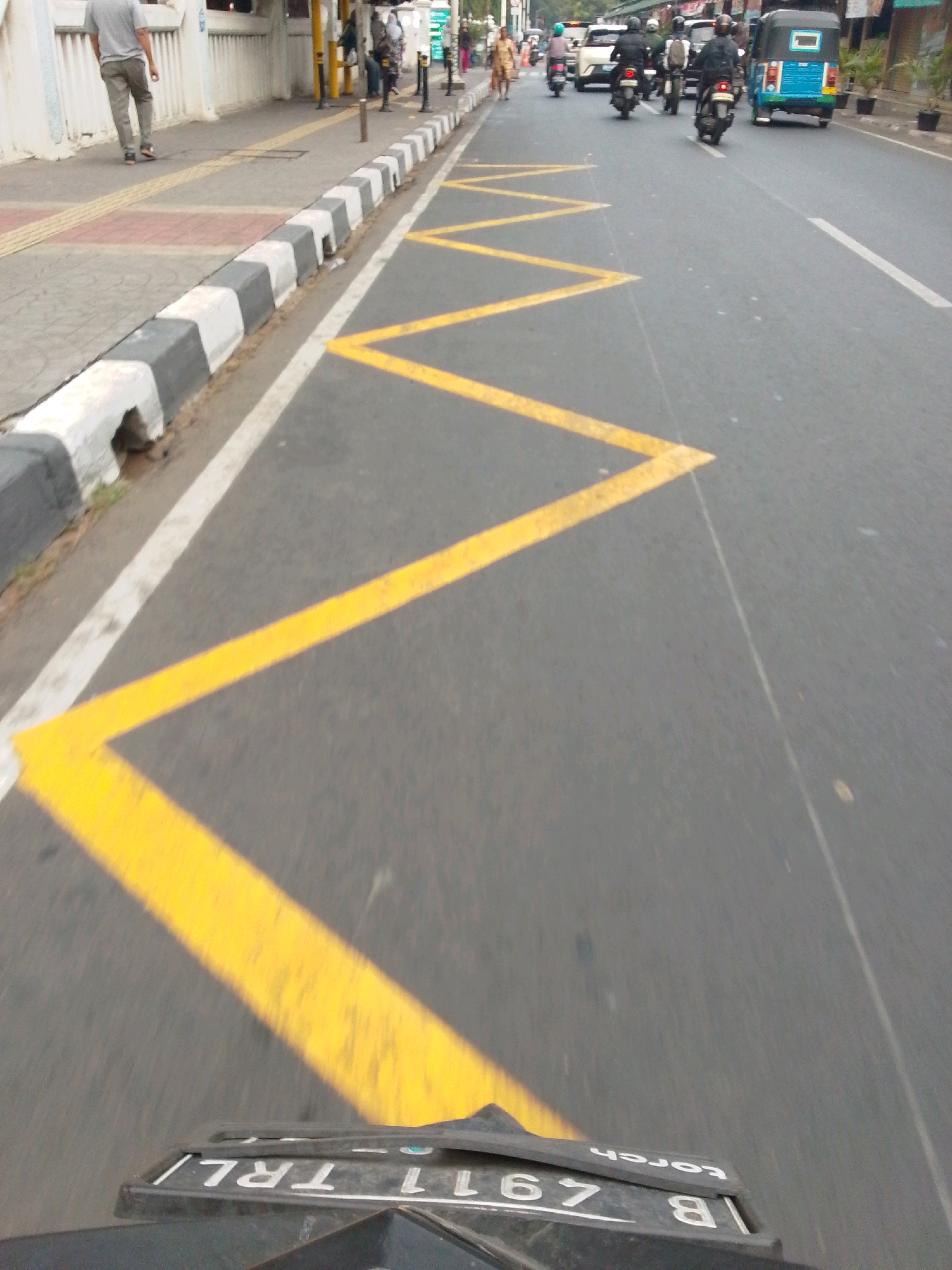 The image shows a road in East Jakarta, Indonesia, with yellow zig-zag markings on the asphalt. A person in a grey shirt and pants walks on a sidewalk next to the road. Several motorcycles and a blue three-wheeled vehicle are visible on the road, some of which are moving. The environment appears to be urban, with buildings and trees in the background. The perspective suggests the image was taken from a moving vehicle. A license plate with the text "B 4911 TRL" and "torch" is visible at the bottom of the image.