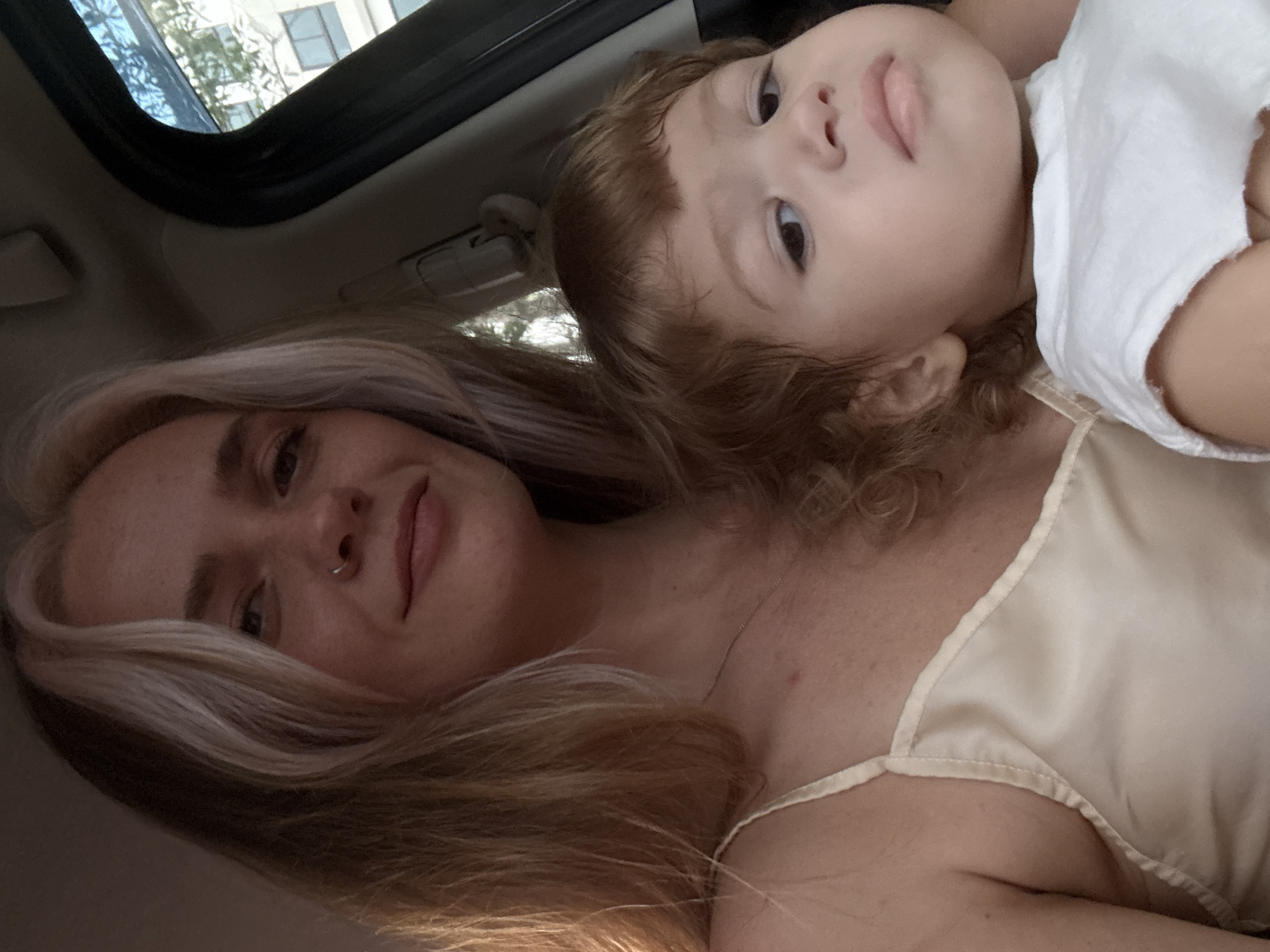 A young woman with blonde hair, featuring streaks of lighter blonde, is holding a toddler. The woman is wearing a pale, silky camisole. The toddler, with curly brown hair, is wearing a white garment. They are likely in the interior of a vehicle, suggested by the car door frame and window visible on the right side of the image. The window shows a glimpse of a building and trees, indicating an urban or suburban outdoor environment. There is no specific detail in the image to place this scene in Choeng Thale, Thailand.