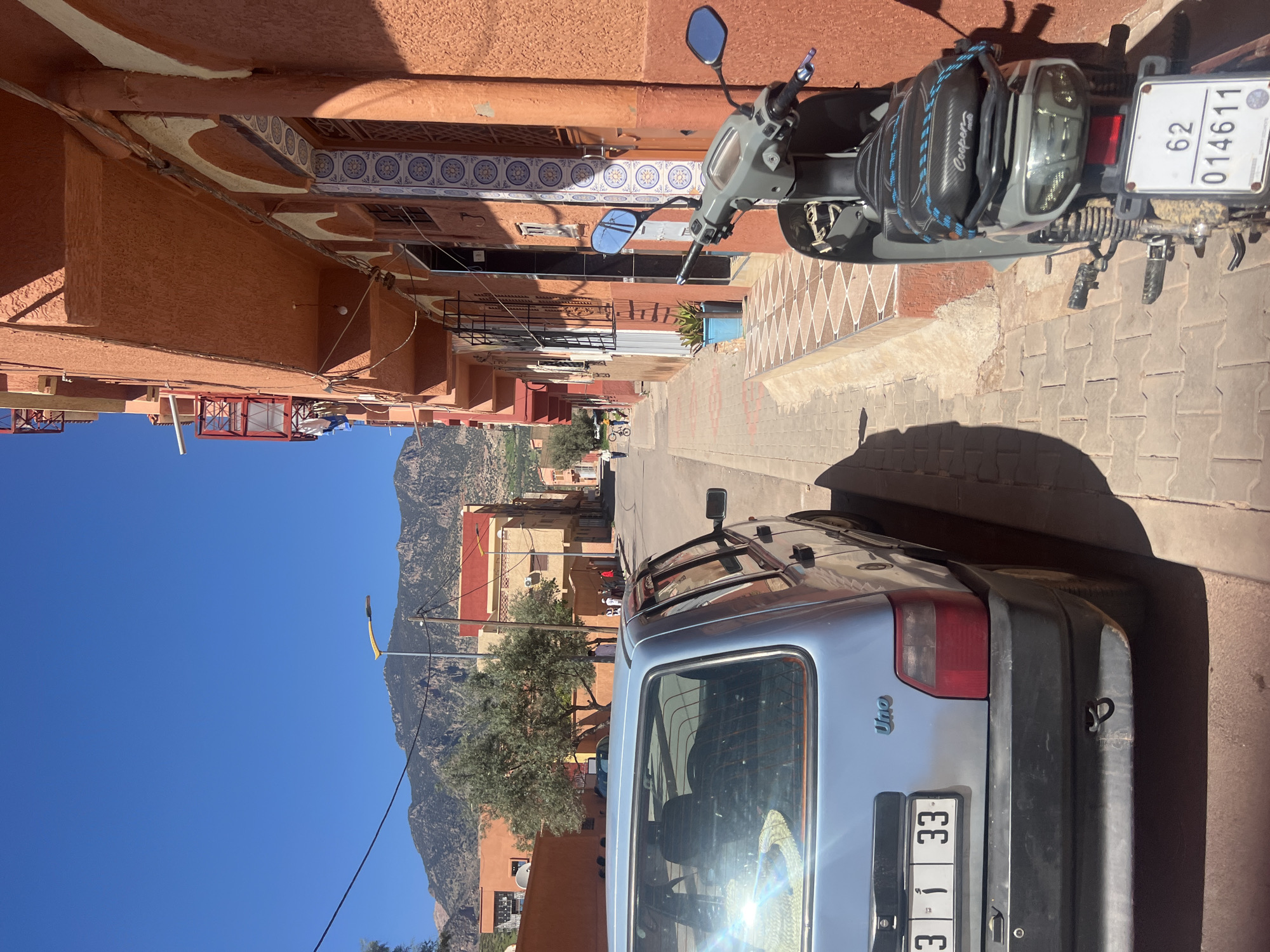 A street scene in Ouaouizert, Morocco, taken during daylight hours. A silver Uno vehicle is parked on the right side of the road. The rear of a scooter with the license plate 62 014611 is visible on the right. The street is paved with interlocking blocks. Buildings with orange facades line both sides of the street. In the distance, mountains are visible under a clear blue sky. There are multiple people scattered throughout the street and some are wearing white clothing.