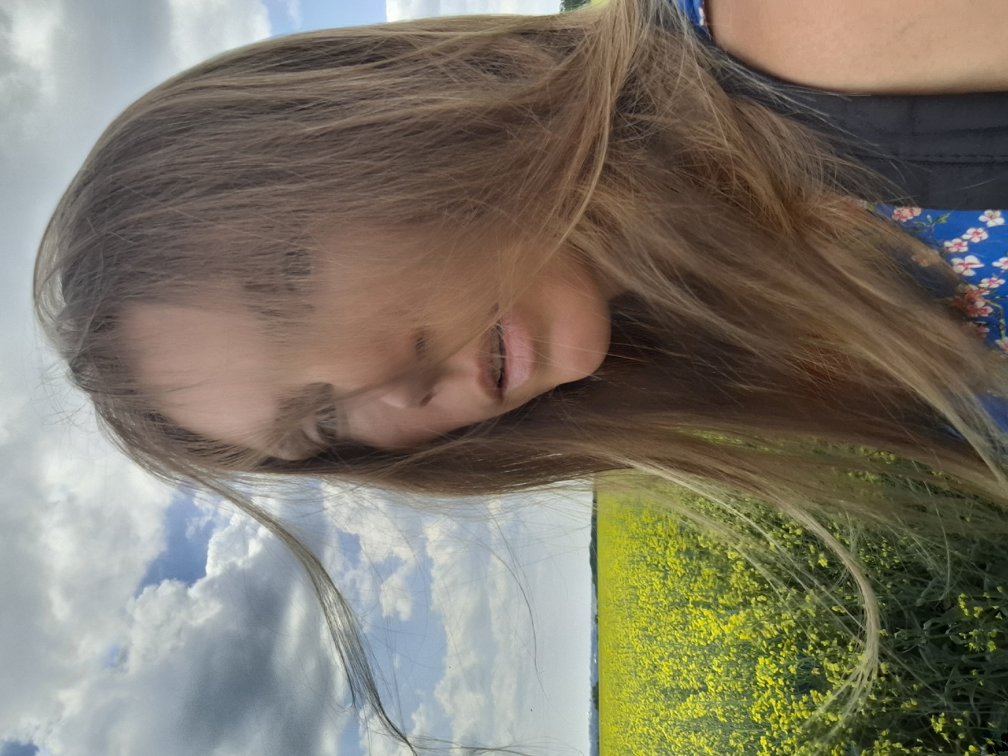 A close-up, medium shot captures a woman with long, blonde hair standing in a field of yellow flowers under a cloudy sky. The woman's face is partially obscured by her hair, and she is looking to the left of the frame. Her mouth is slightly open, and her eyes are not fully visible. She is wearing a blue garment with a floral pattern and a dark backpack. The field of yellow flowers stretches to the horizon, meeting a distant tree line. The sky is a mix of white and grey clouds with patches of blue visible. The image appears to be taken in Treshchevka, Russia, as indicated by the user's prompt.
