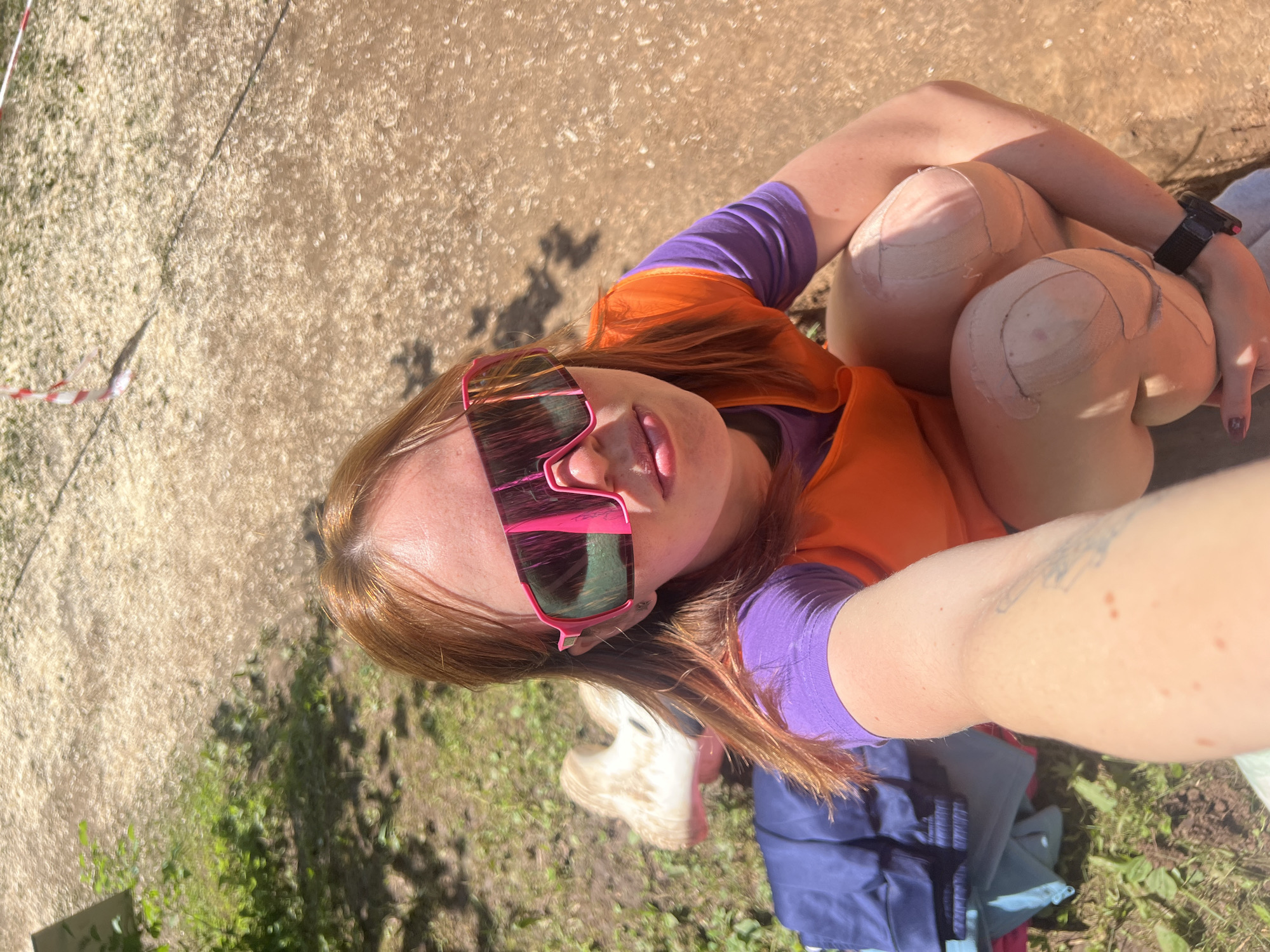 The media file shows a close-up selfie of a woman with reddish-brown hair, wearing pink-framed sunglasses and an orange vest over a purple long-sleeved shirt. Her knees are bandaged, suggesting an injury or athletic activity. The background is a mix of dry, sandy ground and green grass, with some dappled sunlight indicating it's daytime. There are also some dark shadows visible, likely from trees or other objects. A thin red and white striped string or tape is visible in the upper left corner. A blue bag and a white object, possibly a shoe, are partially visible in the lower portion of the image. The overall impression is of an outdoor setting, possibly after an outdoor activity or event, given the bandages and athletic attire.