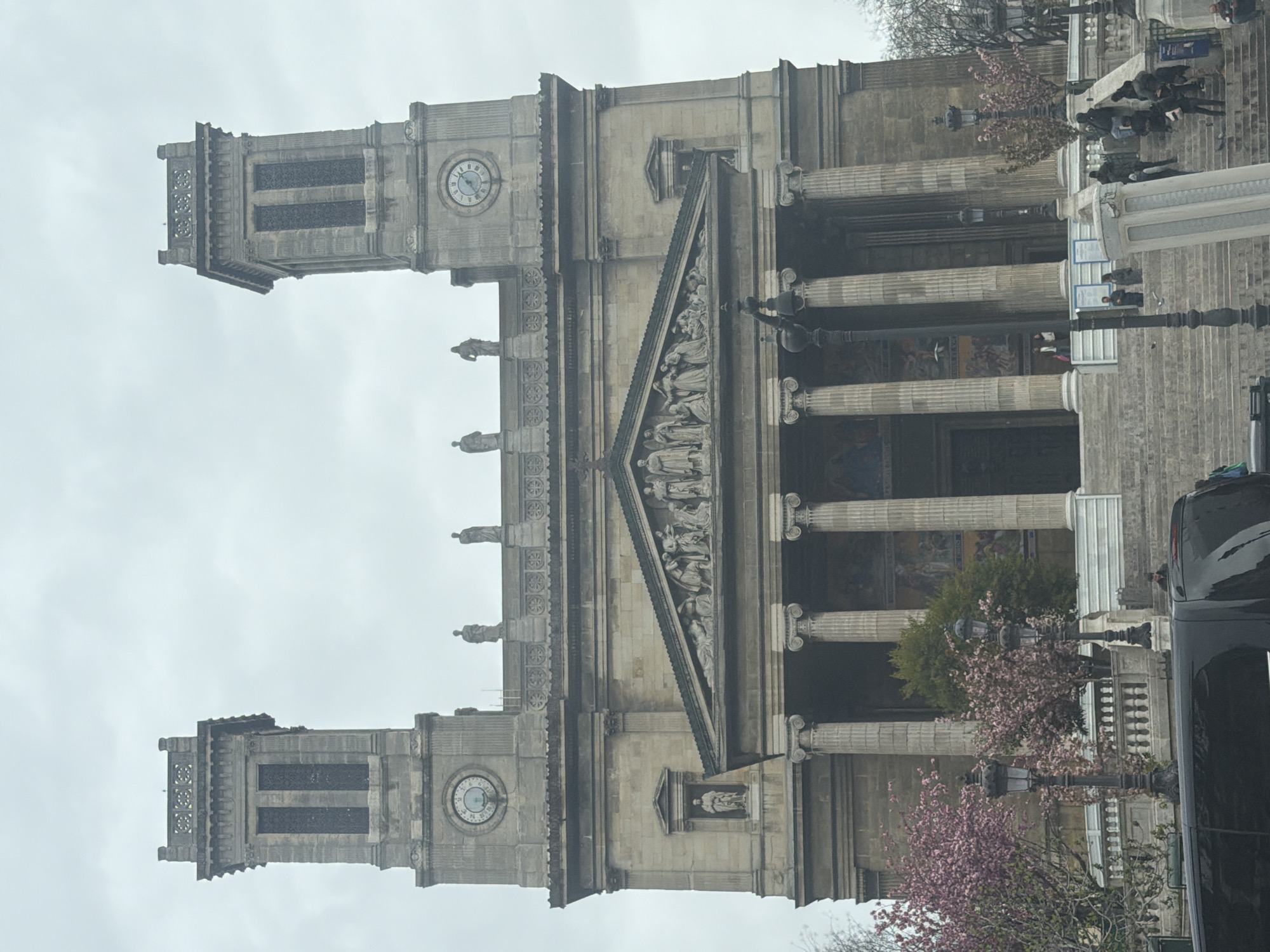 The image showcases the Church of La Madeleine in Paris, France, under an overcast sky. The neoclassical facade, with its prominent Corinthian columns and triangular pediment featuring sculptural reliefs, dominates the scene. Two towers flank the main structure, each equipped with a clock.
The building is approached by a wide set of stairs where indistinct figures of people are present. Street lamps and blossoming trees frame the base of the building. A black car is visible on the right side of the frame, indicating that the photograph was likely taken from street level.