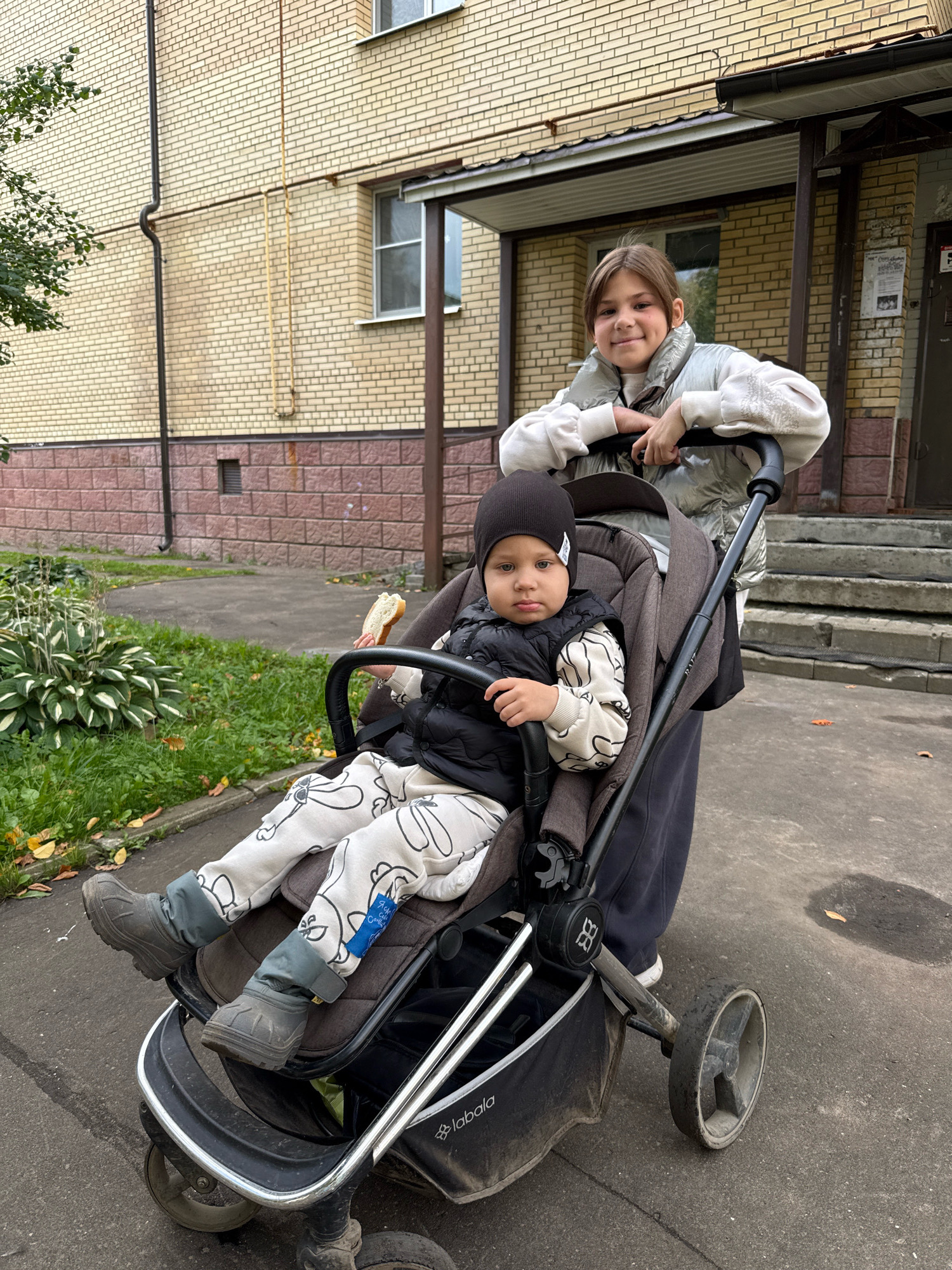 In Rusanovo, Russia, a teenage girl stands behind a baby stroller, her hands resting on its handle. She wears a light-colored puffer vest over a sweatshirt and has a slight smile. A baby is seated in the stroller, facing forward, holding a piece of bread. The baby is dressed in a dark vest and a patterned outfit, with a dark beanie on their head. The stroller is a dark grey color with a silver frame and black wheels. It sits on a concrete sidewalk. Behind them is a light-colored brick building with a brown entrance and steps. Green bushes and scattered fallen leaves are visible to the left of the building. The lighting suggests an overcast day.