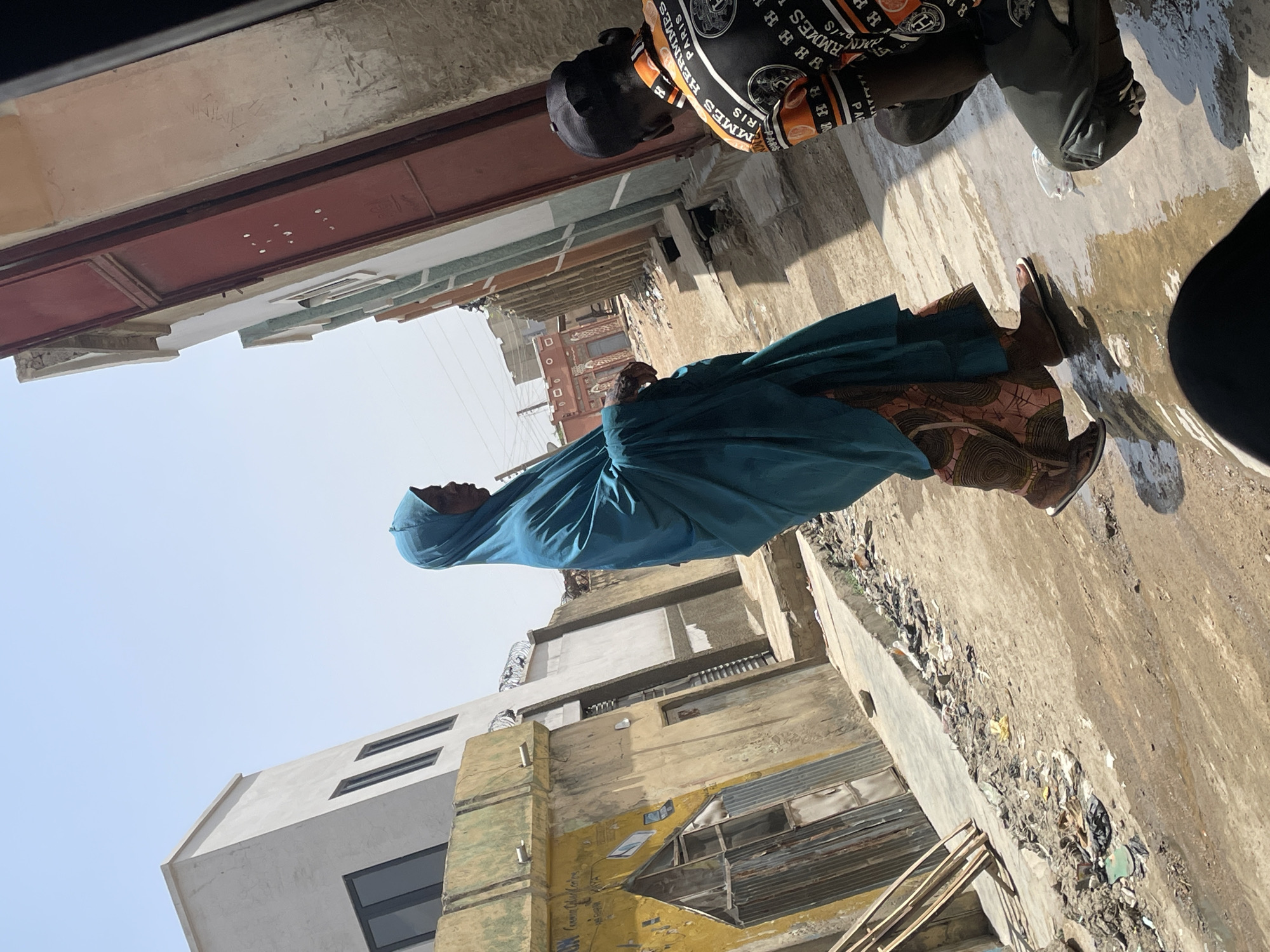 A street scene in the city of Unknown, Unknown shows a woman wearing a blue head covering and a patterned garment walking along a street next to a man wearing a black cap and shirt with an orange and white pattern. Buildings of varying heights and colors line the street. The ground is wet and appears to have some trash and debris scattered along the edges. The sky is pale blue.