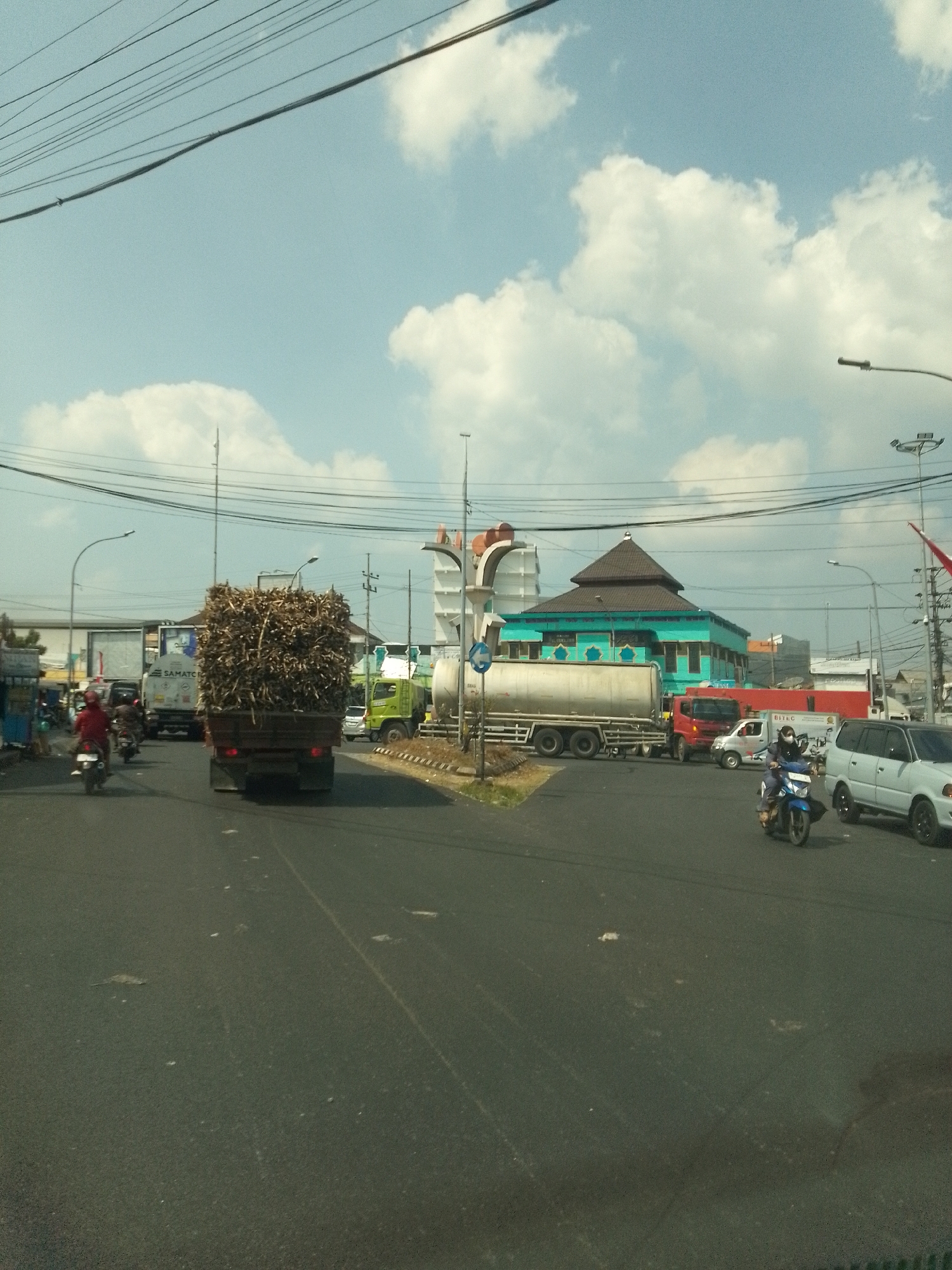This is a mid-range, eye-level, outdoor, daytime, horizontal, color, medium-resolution, slightly blurry, overexposed, eye-level, full shot of a busy intersection in Nguwok, Indonesia. The sky is a bright blue with scattered white clouds. Power lines crisscross the upper part of the frame. On the road, several vehicles are present, including a large truck carrying a load of dried sugarcane stalks in the foreground. A green truck is to the right of the sugarcane truck, and a white tanker truck is further back. Several motorcycles are also visible, with riders wearing helmets. A light-blue building with a multi-tiered brown roof, possibly a mosque, is located in the background. The road surface is asphalt and appears to be in good condition, with visible lane markings. The overall scene depicts a typical day at a busy intersection in an Indonesian town.