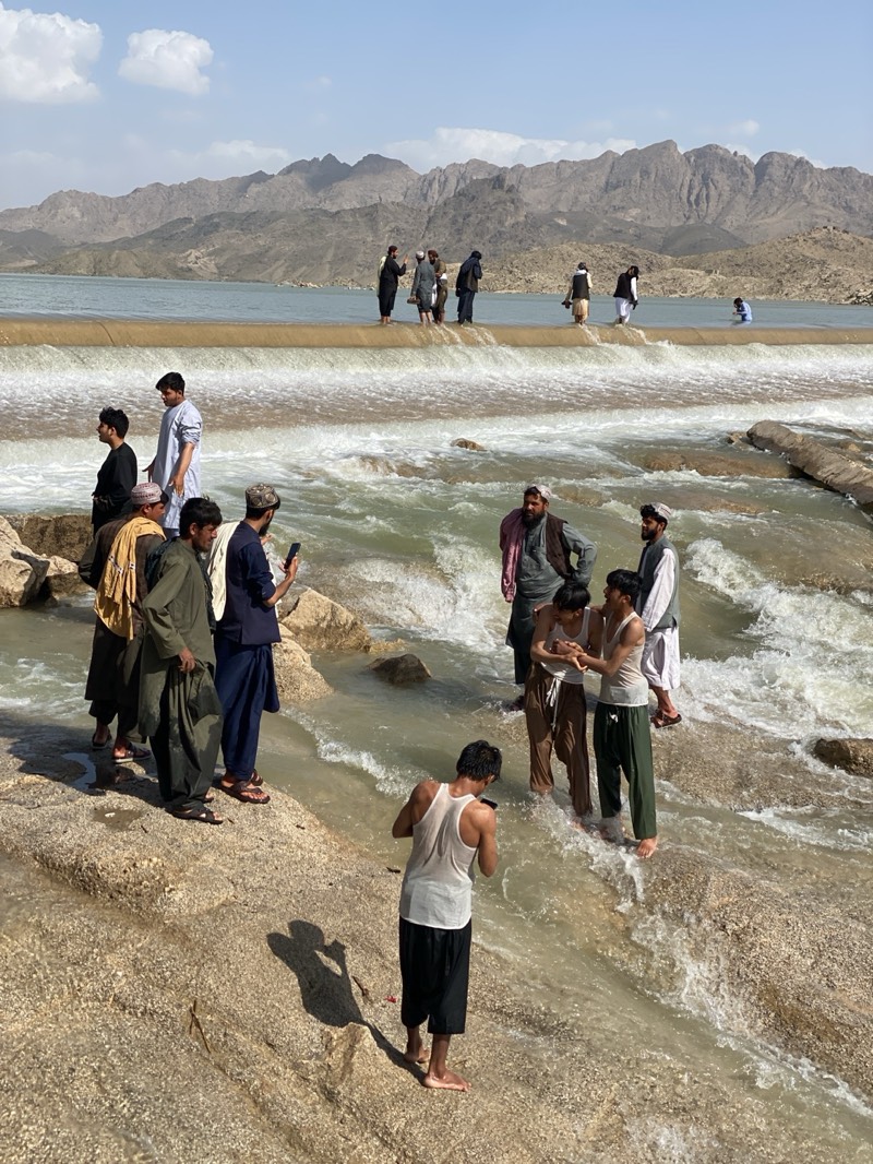 A group of people are gathered at a dam and spillway with a river. Some of the people are standing on the dam, while others are in the river. The people in the river are interacting with each other. The river is flowing rapidly and has whitewater rapids. The background shows a mountain range. The sky is clear with a few scattered clouds. The location is near the city of Kim in Afghanistan.