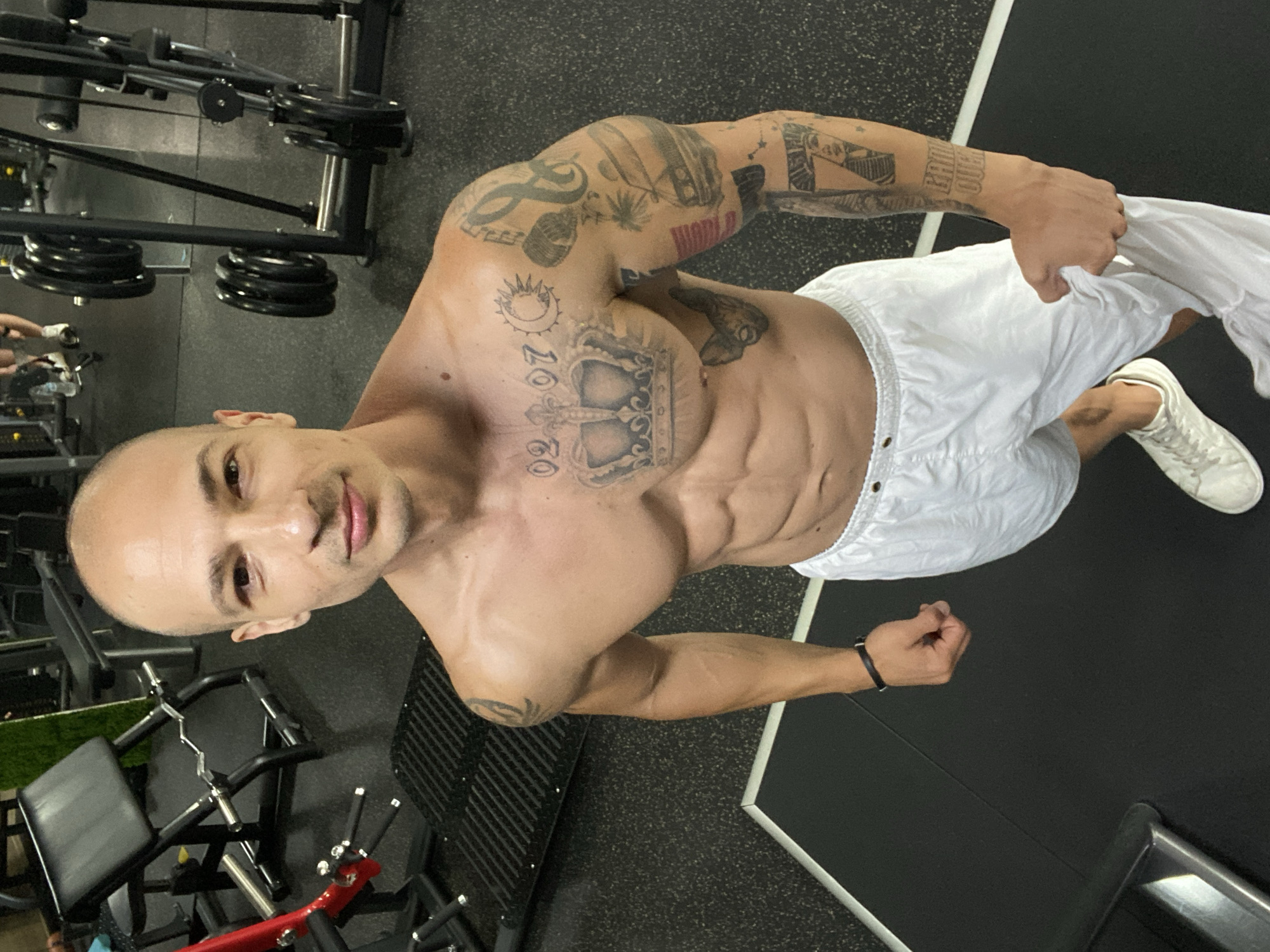 A bald, shirtless man with prominent abdominal muscles and tattoos on his chest and arms stands in a gym. He is wearing white shorts and white sneakers. He is holding a white towel in his right hand. In the background, there are various weightlifting machines and equipment. The flooring appears to be black rubber mats. The lighting suggests an indoor environment, likely a gym. The scene is photographed from a high-angle perspective, looking down at the man.