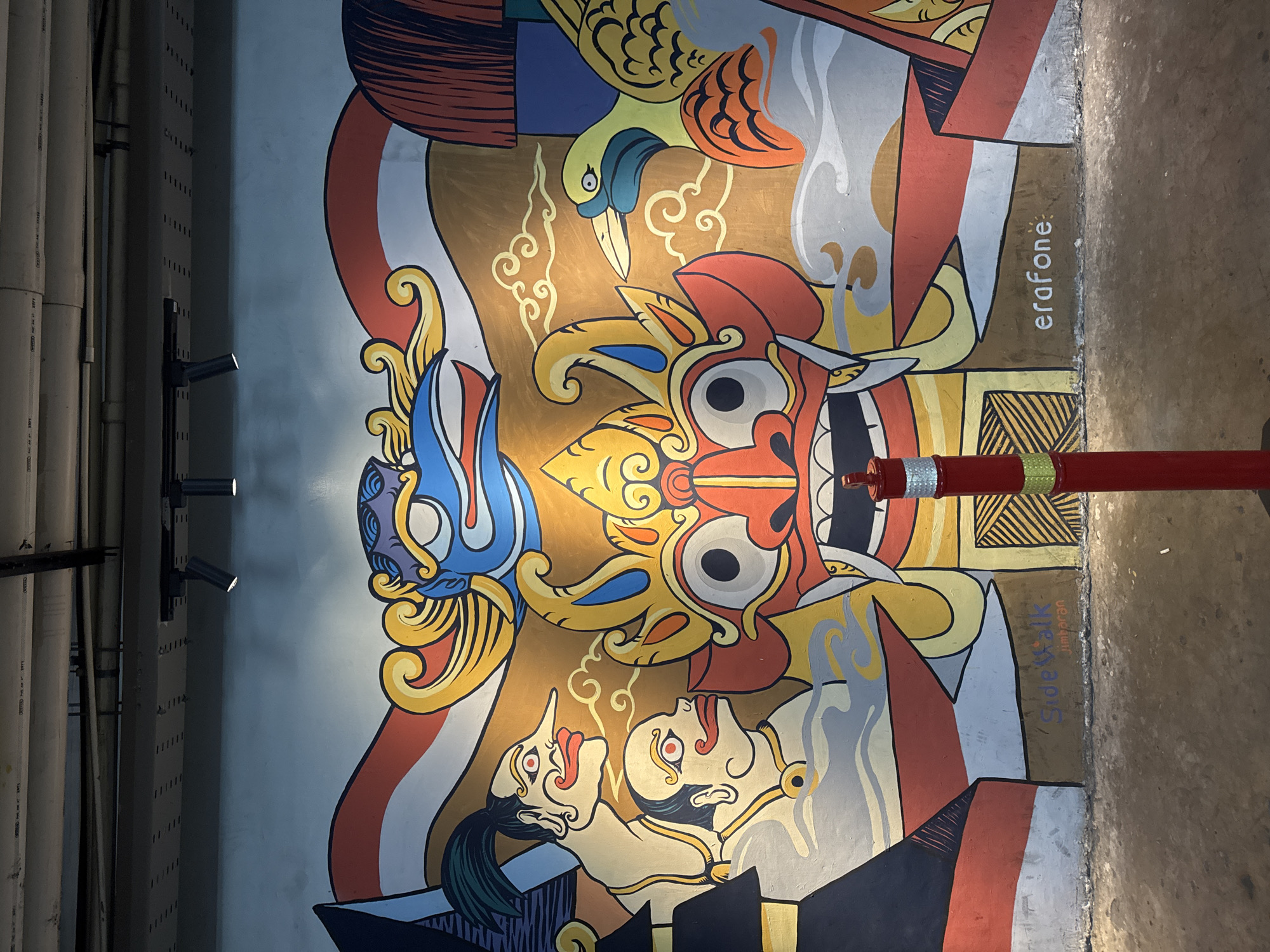 The image captures a vibrant mural on a wall in Kuta Selatan, Indonesia, likely an outdoor or semi-outdoor public space given the ground texture and overhead piping. The mural depicts traditional Indonesian motifs with bold colors and stylized figures. Dominating the center is a large, fierce-looking mask with prominent red lips and large eyes, rendered in yellow, red, and blue. Flanking this central figure are depictions of two human faces with distinctive red-lipped smiles and red eyes, one with a long, pointed nose and a green topknot, and another with a shaved head. To the right, a yellow bird with blue and green accents is shown, possibly a cockatoo or similar tropical species. The background features wavy lines suggesting clouds or smoke, set against a golden backdrop, and is framed by horizontal bands of red and white, reminiscent of the Indonesian flag. In the lower portion of the mural, the text "Sidewalk Jimbaran" is visible in purple and white, and to the right, "erafone" is written in white. A bright red bollard with reflective silver and green bands stands prominently in the foreground, obscuring a small portion of the mural and the ground. The ground appears to be a mix of concrete and dirt, with some debris visible. Overhead, electrical conduits and lighting fixtures are mounted on the ceiling or upper wall.