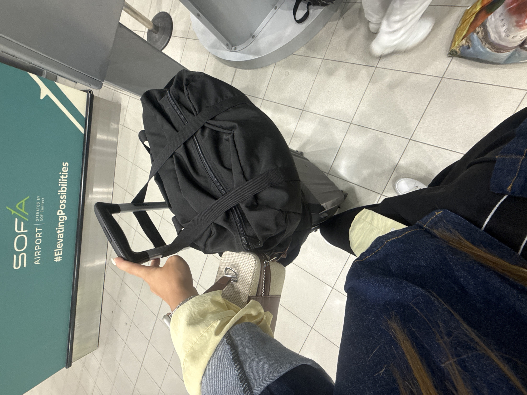 The image is a first-person perspective shot taken in an airport in Sofia, Bulgaria. In the foreground, a person's hand, with neatly manicured fingernails, is seen holding the handle of a black rolling suitcase. A black duffel bag is resting on top of the suitcase, secured by straps. To the left of the suitcase, a small, light-colored toiletry bag with brown accents is visible. The person is wearing a light yellow jacket over a denim garment, and dark pants. A portion of the airport's tiled floor is visible, along with a large teal-colored advertisement board in the background bearing the text "SOFIA AIRPORT" and "#ElevatingPossibilities". To the right, a part of an airport counter or structure is visible.