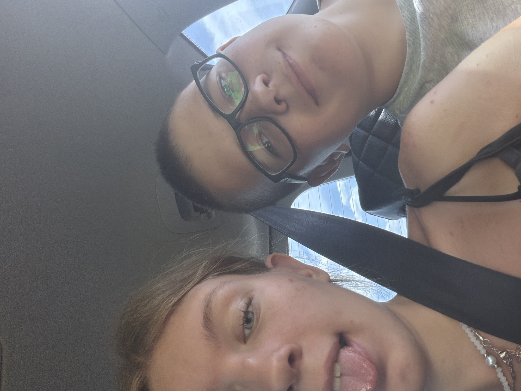 In the city of Novoe Feizullovo, Russia, two individuals are captured in a selfie taken from within a vehicle. The person on the left, a young female with blonde hair, is positioned closer to the camera, her face partially visible as she sticks her tongue out. She is wearing a necklace with pearl and metallic charms and appears to have a seatbelt across her body. To the right, a young male with short hair and glasses is also visible. He is looking towards the camera with a slight smile. The interior of the car is discernible, with a dark-colored car seat visible behind the male, and a glimpse of the sky and clouds through the rear window. The lighting suggests it is daytime.