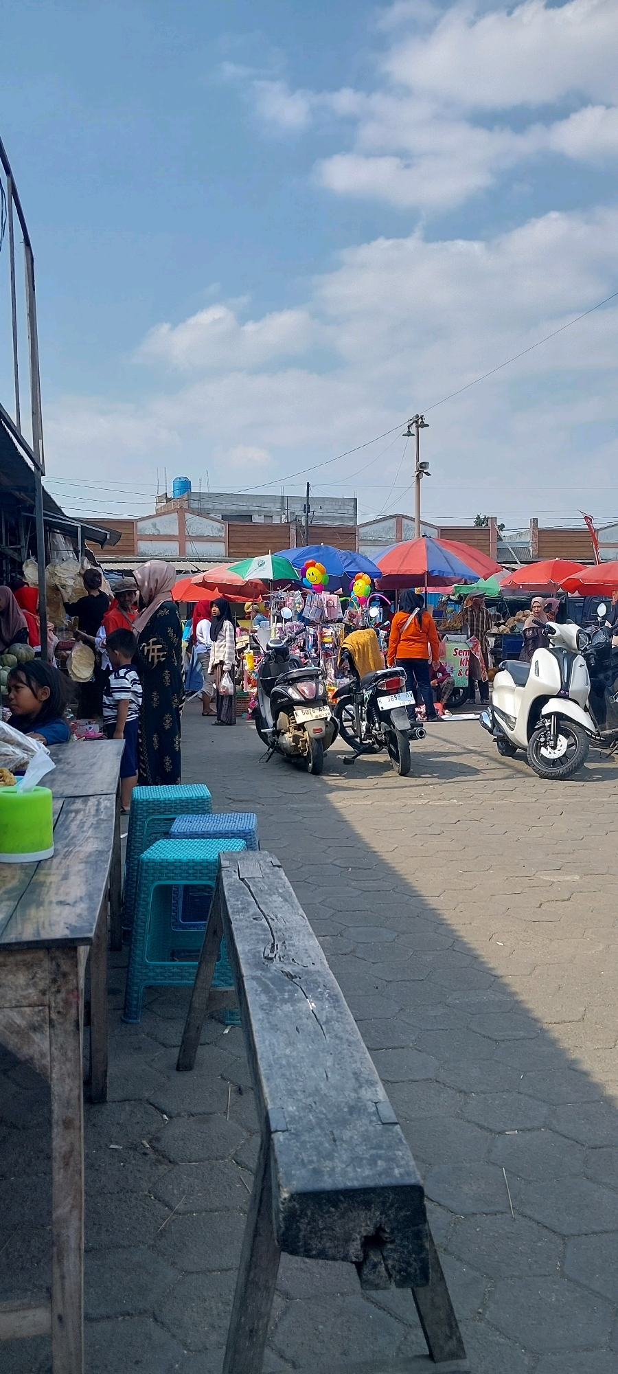 This is a vertical, eye-level outdoor photograph of a bustling market scene in Pecangaan, Indonesia. The sky is a bright blue with scattered white clouds. In the foreground, to the left, a rustic wooden table is visible, with a bright green plastic container on it. Three colorful plastic stools, two turquoise and one blue, are arranged in a row behind the table.
The middle ground of the image depicts a lively market with numerous stalls and people. Several motorcycles are parked in the paved walkway. Various vendors are seen attending to their stalls, which are covered by colorful umbrellas, including red, blue, and green ones. One stall displays children's toys, including balloons with smiley faces. People are browsing and interacting, dressed in casual clothing, some wearing headscarves.
In the background, buildings with tiled roofs and a blue water tank are visible. The ground is paved with hexagonal stones. The overall impression is of a vibrant and active marketplace under a clear sky.