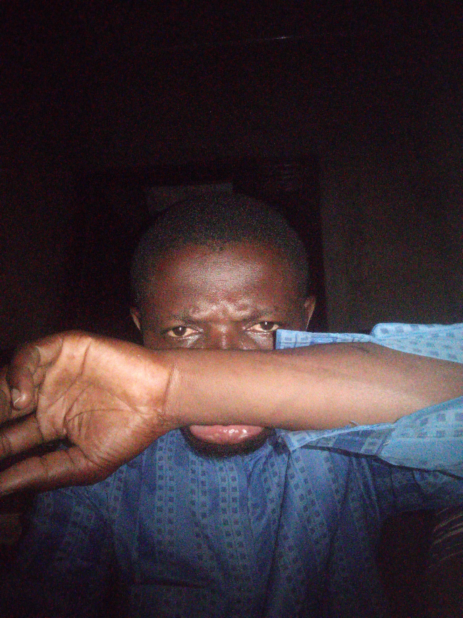 A close-up, dimly lit, indoor shot shows a Black individual's face and arm. The person's left arm is bent across their face, with their hand obscuring their mouth and nose, and resting against their cheek. One eye is visible, looking directly at the camera. The person is wearing a blue shirt with a repeating dark blue pattern. The background is dark and indistinct, suggesting an indoor setting with low lighting. The image appears to have been taken with a low-quality camera, resulting in grainy texture and muted colors. The location in Tempure, Nigeria, is not discernible from the visual information provided in the image.