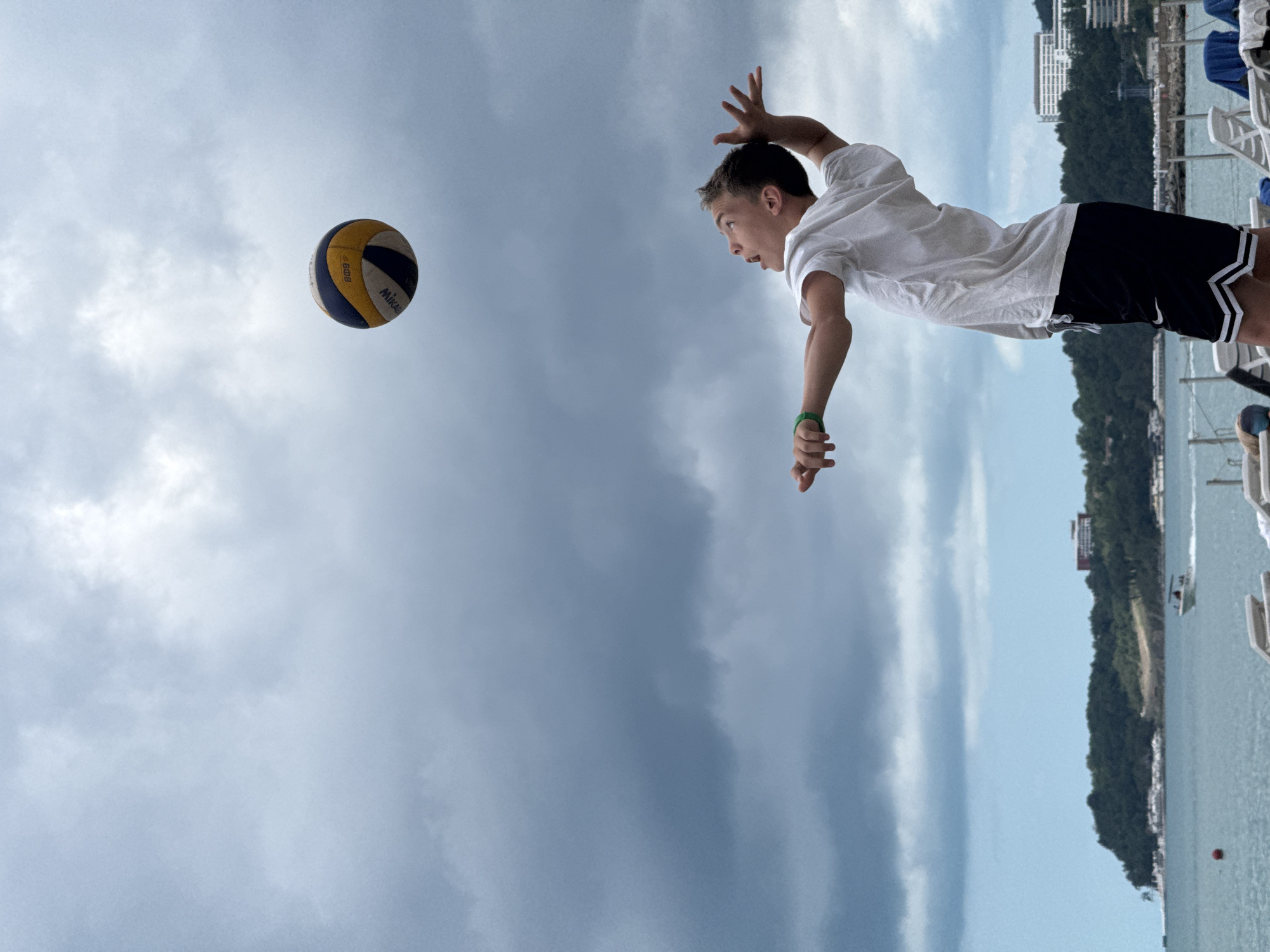 A young boy in a white t-shirt and black shorts with white stripes on the hem is playing volleyball on a beach. He is in the process of hitting the ball, which is in the air above him. The boy's arms are extended, and his face shows concentration. The background features a cloudy sky, the sea, a coastline with trees and buildings, and lounge chairs on the sand. The location is identified as the city of Nebug, Russia.