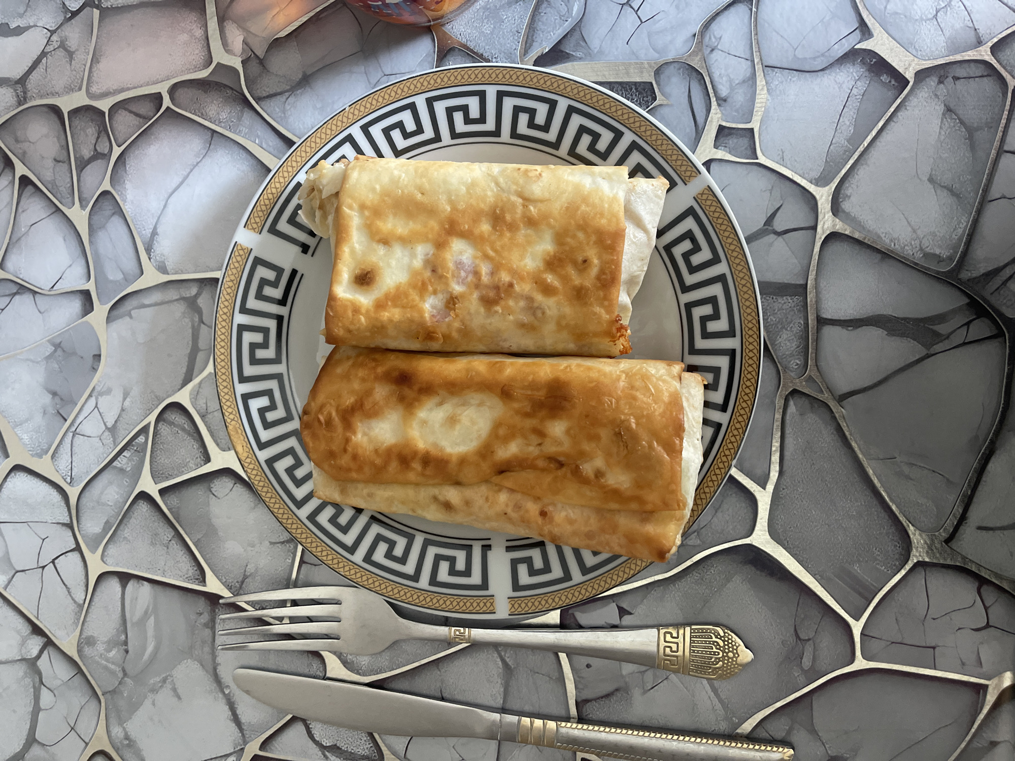 Two golden-brown, rectangular wraps are presented on a white plate with a decorative black and gold rim. The wraps appear to be filled and lightly fried, with some visible filling peeking out from the edges of one wrap. To the left of the plate are a silver knife and fork, both featuring ornate gold handles with intricate geometric patterns. The plate and utensils are placed on a grey surface with a striking, modern design resembling cracked stone or abstract geometric shapes with metallic silver and black accents. The background exhibits a pattern consistent with the table surface. The scene is captured from a top-down perspective. This image was taken in Novxanı, Azerbaijan.