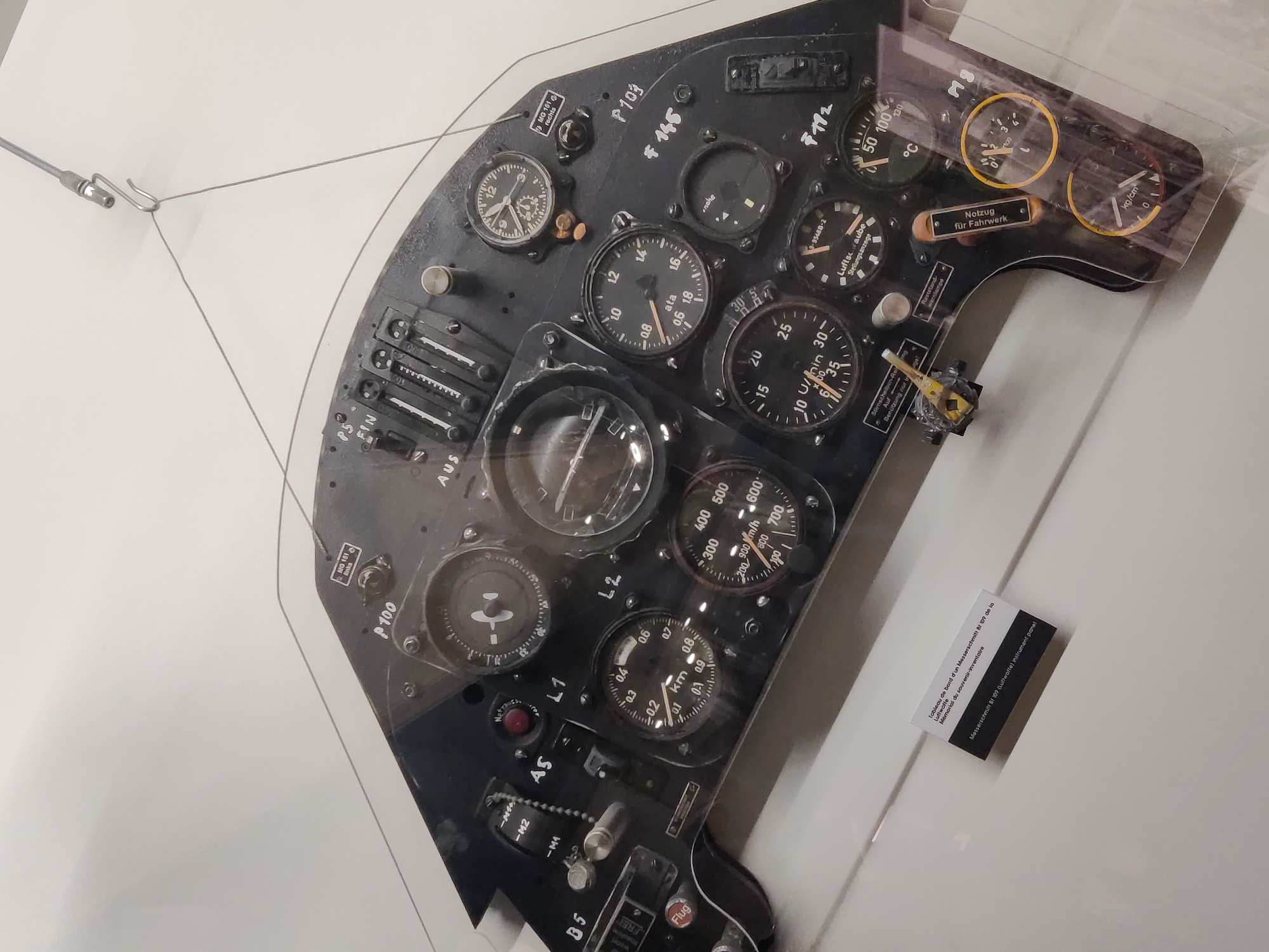 The image shows a close-up view of an airplane's instrument panel, likely a Messerschmitt Bf 109, as indicated by a label in the lower left corner. The panel is black and filled with various gauges, dials, and switches. Some of the visible instruments include a clock, a tachometer (U/min), an altimeter, a variometer (indicated by vertical markings), and a thermometer (in °C). There are also labels in German on some of the components. The instrument panel is housed within a clear protective casing, and the overall setting appears to be a museum or exhibition, suggested by the diffused lighting and the clean, white background. A small card with descriptive text in French and English, identifying the panel as being from a Messerschmitt Bf 109, is placed in front of the casing. There are no people visible in the image. The scene does not provide any specific information that would allow for the determination of its location in the city of Dunkirk, France.