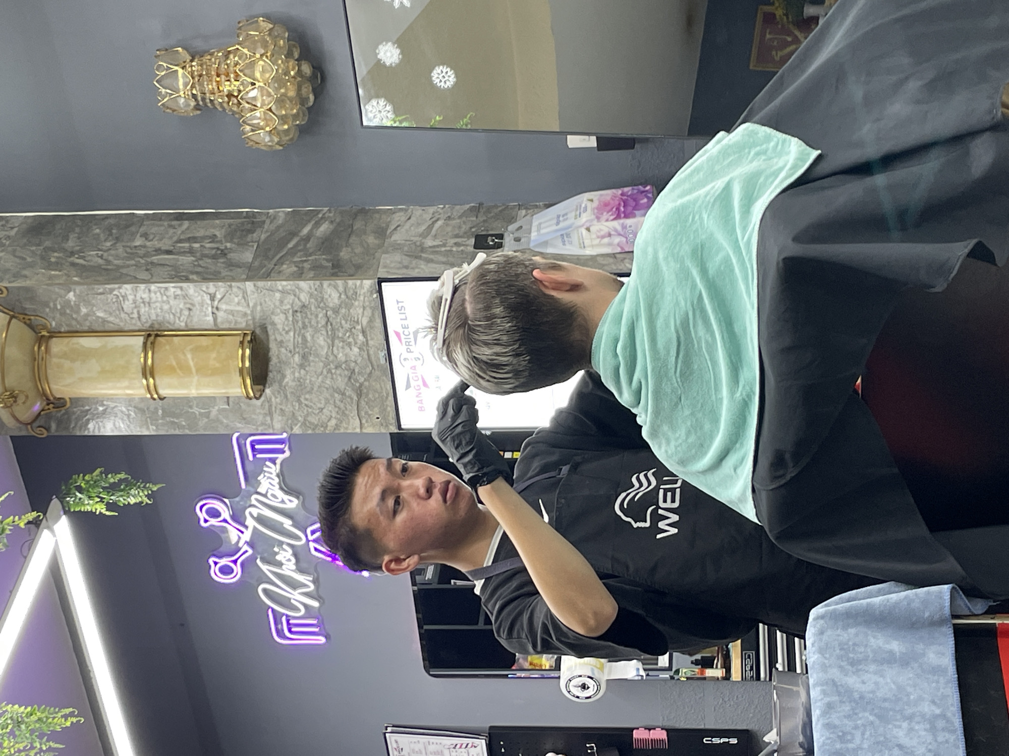 This image depicts a barber in Xuân Hương, Đà Lạt, Vietnam, performing a haircut on a client. The barber, a young East Asian male wearing a black apron with the "WELL" logo, is focused on trimming the client's hair. The client, seen from behind, has short, light-colored hair and is draped in a teal towel.
The barbershop environment is visible, with a neon sign in the background featuring scissors and Vietnamese text. The walls are painted a dark gray, and a decorative golden chandelier hangs on the right side of the frame. A price list is displayed on a board to the left of the client. Various barber tools and equipment are subtly visible on shelves and surfaces, suggesting a functional barbershop setting. The lighting appears to be a mix of overhead lights and ambient light from the neon sign.