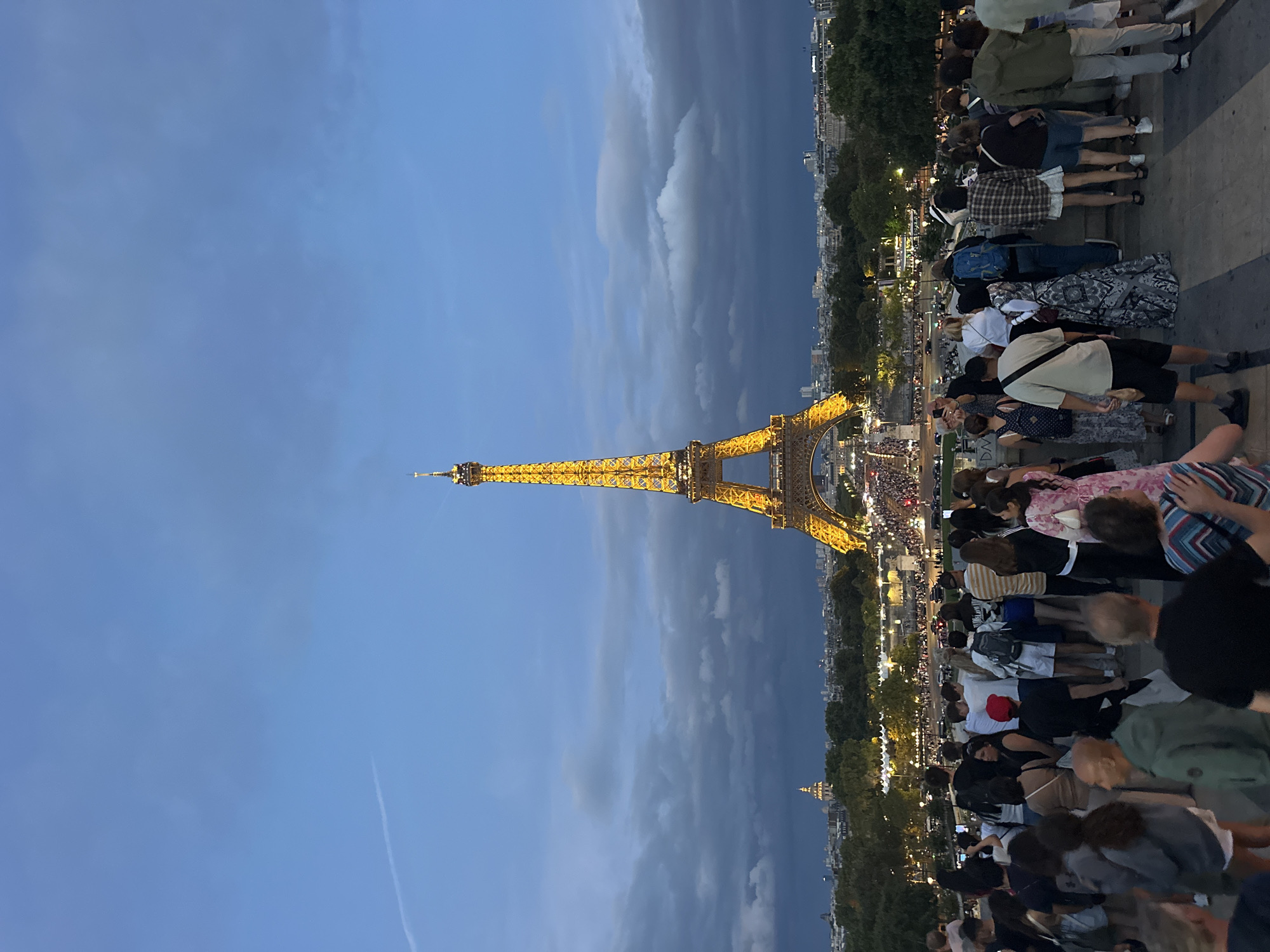 This is a nighttime, full-shot, eye-level, outdoor photograph of the Eiffel Tower in Paris, France. The iconic landmark is illuminated with golden lights and stands prominently in the center of the frame, against a dark blue twilight sky with some clouds. A thin white contrail is visible in the upper left quadrant of the sky. A crowd of people are gathered in the foreground, looking towards the tower and the cityscape behind it. They are standing on a raised platform or walkway, with the city lights and trees extending into the middle ground. The crowd appears diverse in age and attire, with individuals and small groups visible. Some are taking pictures with their phones, while others are simply observing the view. The overall atmosphere is one of tourism and admiration of the landmark.