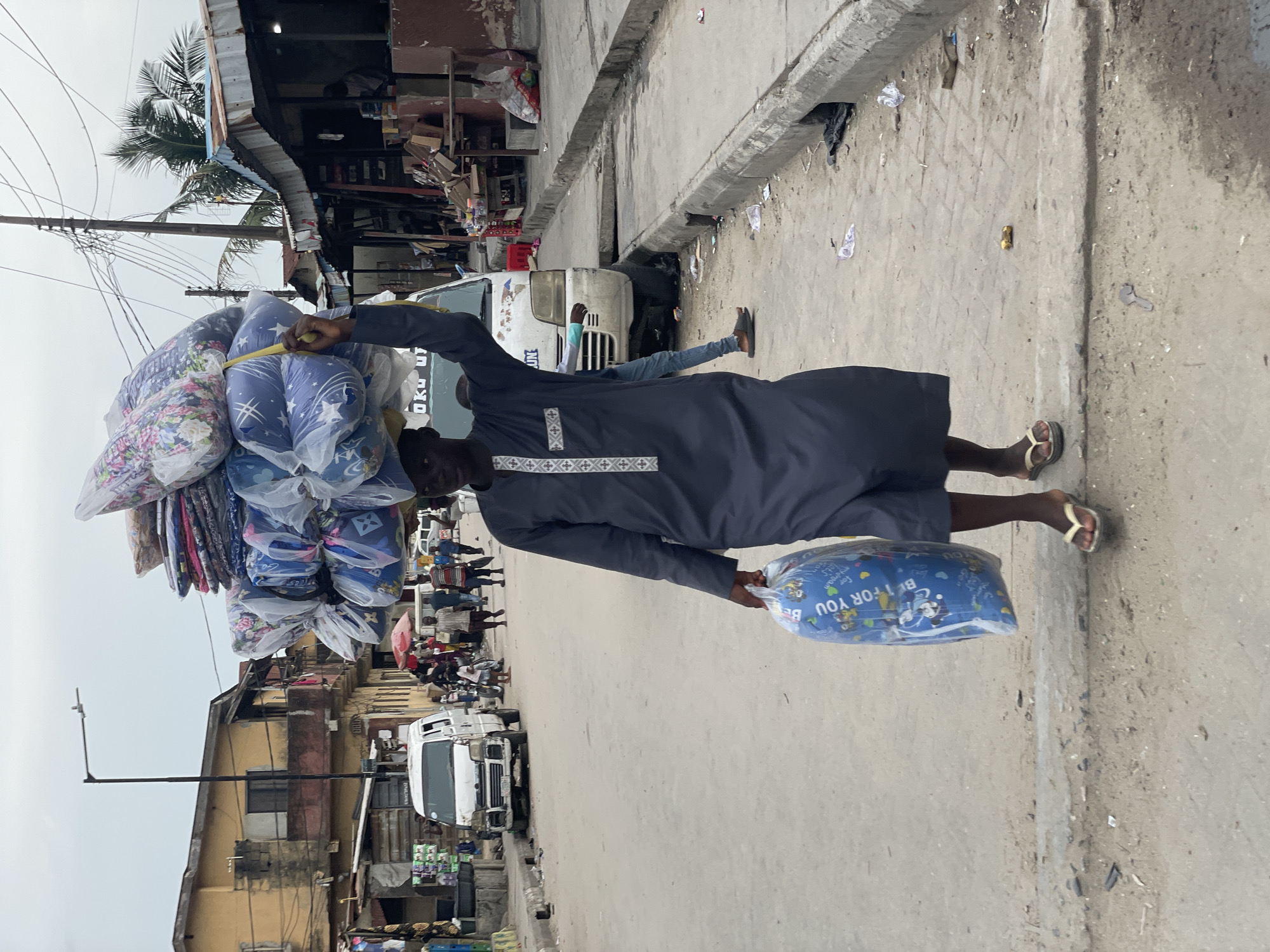 The image captures a man walking down a bustling street, likely a market area in Ado, Nigeria. He is the central figure, expertly balancing a large, tall stack of merchandise on his head. The items are individually wrapped in clear plastic, appearing to be pillows, bedding, or cushions, with some packages featuring floral designs and others a blue pattern with stars, moons, and cartoon characters. In his left hand, he carries an additional blue, rectangular package, also with cartoon characters and the visible text "FOR YOU".
The man wears a dark blue or grey traditional tunic, possibly an agbada, adorned with white embroidered patterns on the chest and lower hem, and is wearing flip-flops.
The street is concrete, with a raised sidewalk to the right. The ground is somewhat unkempt, with scattered debris such as plastic bags and other litter. Along the left side of the street, there are several buildings, appearing somewhat aged and possibly commercial or residential, with corrugated metal roofs. Numerous power lines are strung overhead, and a tall street light pole stands on the left.
In the background, a white van is parked, with the letters "UN" partially visible on its front. Another white vehicle is seen further down the road, and several other people are visible, indicating a busy street scene. The lighting suggests it is daytime, possibly under an overcast sky, creating a soft, diffused light. The overall impression is one of everyday commerce and active street life in an urban setting.