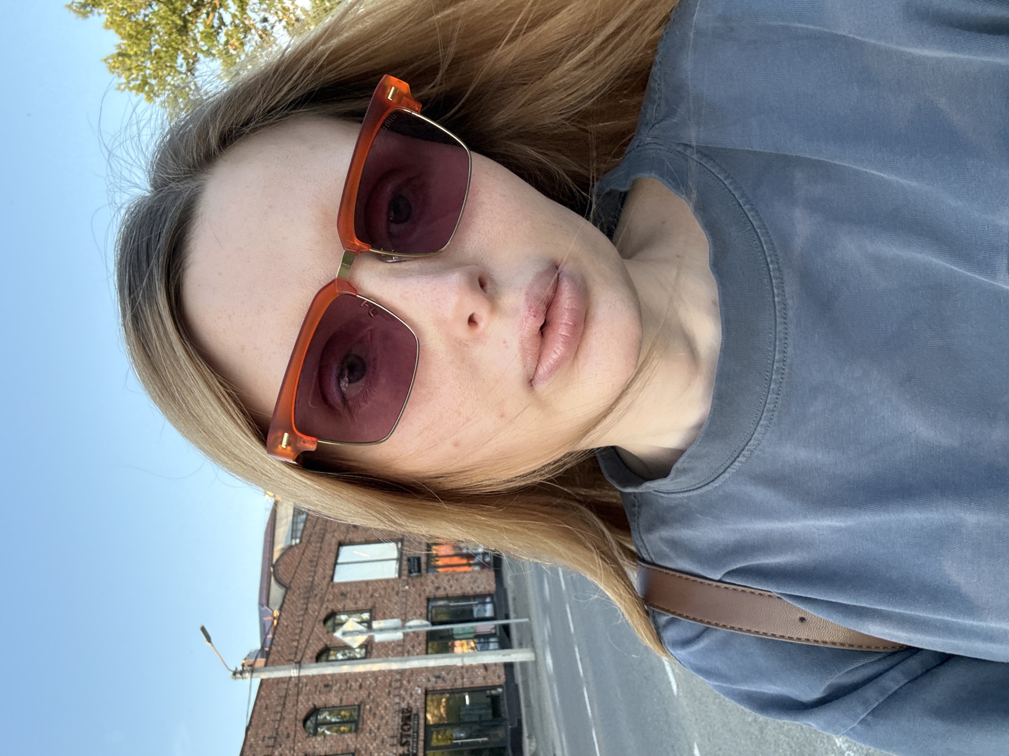 A young woman is taking a selfie outdoors. She is wearing large, orange-framed sunglasses and a grey t-shirt. Her blonde hair is long and down, framing her face. The background shows a blue sky, trees, and a brick building with multiple windows. A street and a streetlight are also visible. The lighting suggests it is daytime, possibly late afternoon given the angle of the sun. The woman's expression is neutral. There is some visible text on the building, partially obscured, that appears to be "STORE".