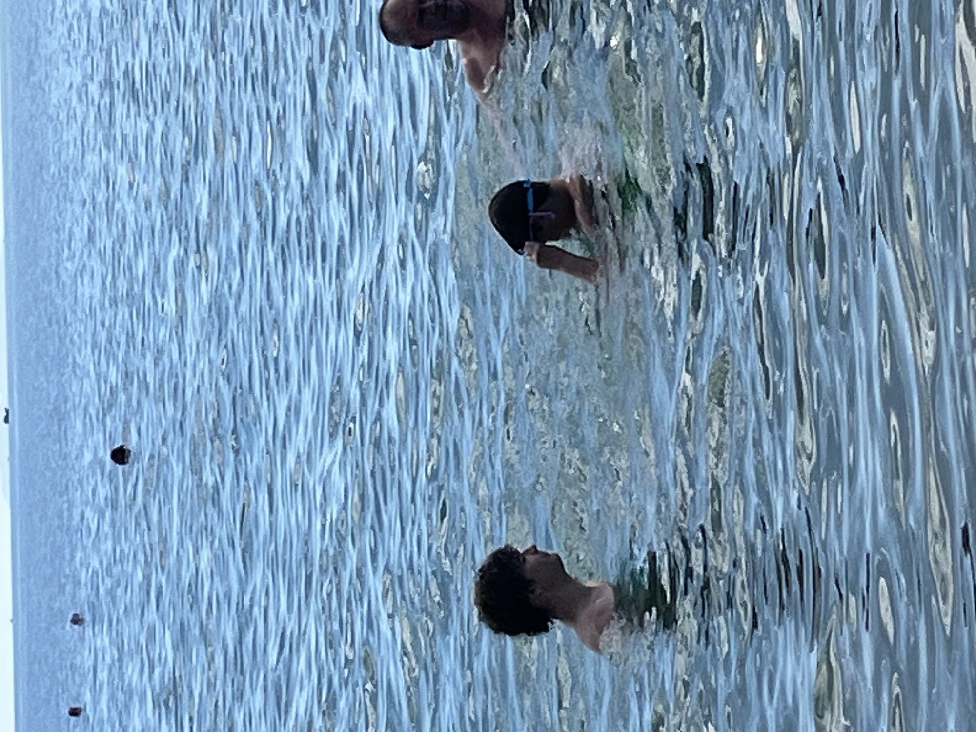 In Nha Trang, Vietnam, a body of calm, rippling blue water stretches towards a hazy horizon, dotted with distant buoys and a faint shoreline. In the foreground, three individuals are partially submerged. To the left, a young person with dark, curly hair is seen in profile, looking towards the right, their shoulders and upper chest visible above the water. In the center, another young person, wearing swimming goggles perched on their forehead, is looking towards their left. To the far right, an adult male with short, bald hair and a relaxed posture is looking towards the left. The water is moderately choppy, with sunlight reflecting off the surface, creating a bright, airy atmosphere.