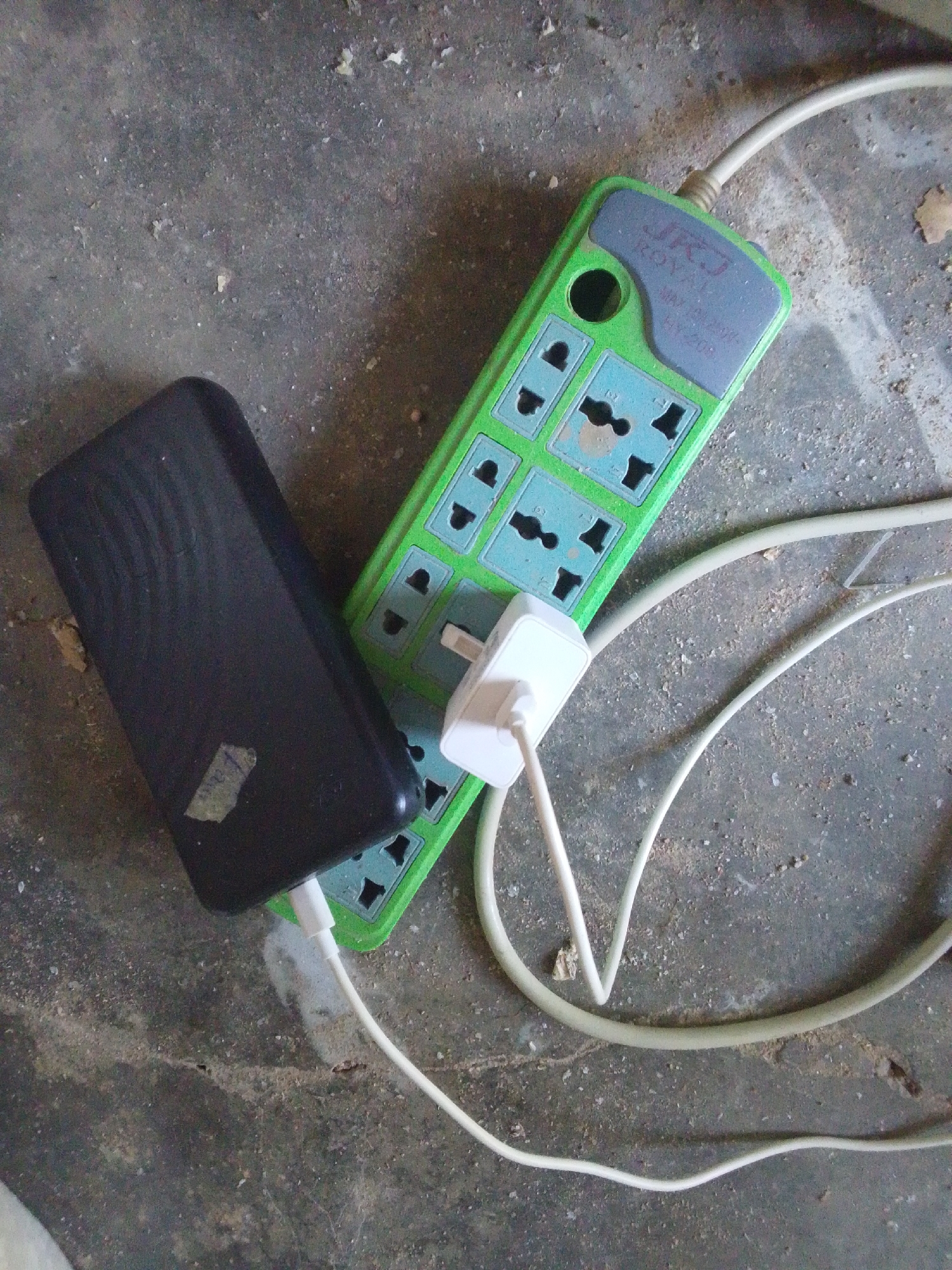 An overhead shot captures a black power bank and a green surge protector lying on a dusty, concrete surface. The surge protector, labeled "JRJ ROYAL 100-250V 10A", has multiple universal outlets and a white adapter plugged into one of them. A white cable extends from the adapter and connects to the black power bank. The environment suggests an outdoor or unfinished indoor space, potentially in Jalingo, Nigeria, given the rough texture of the ground and the presence of debris. There are no people visible in the image, and the scene appears static.