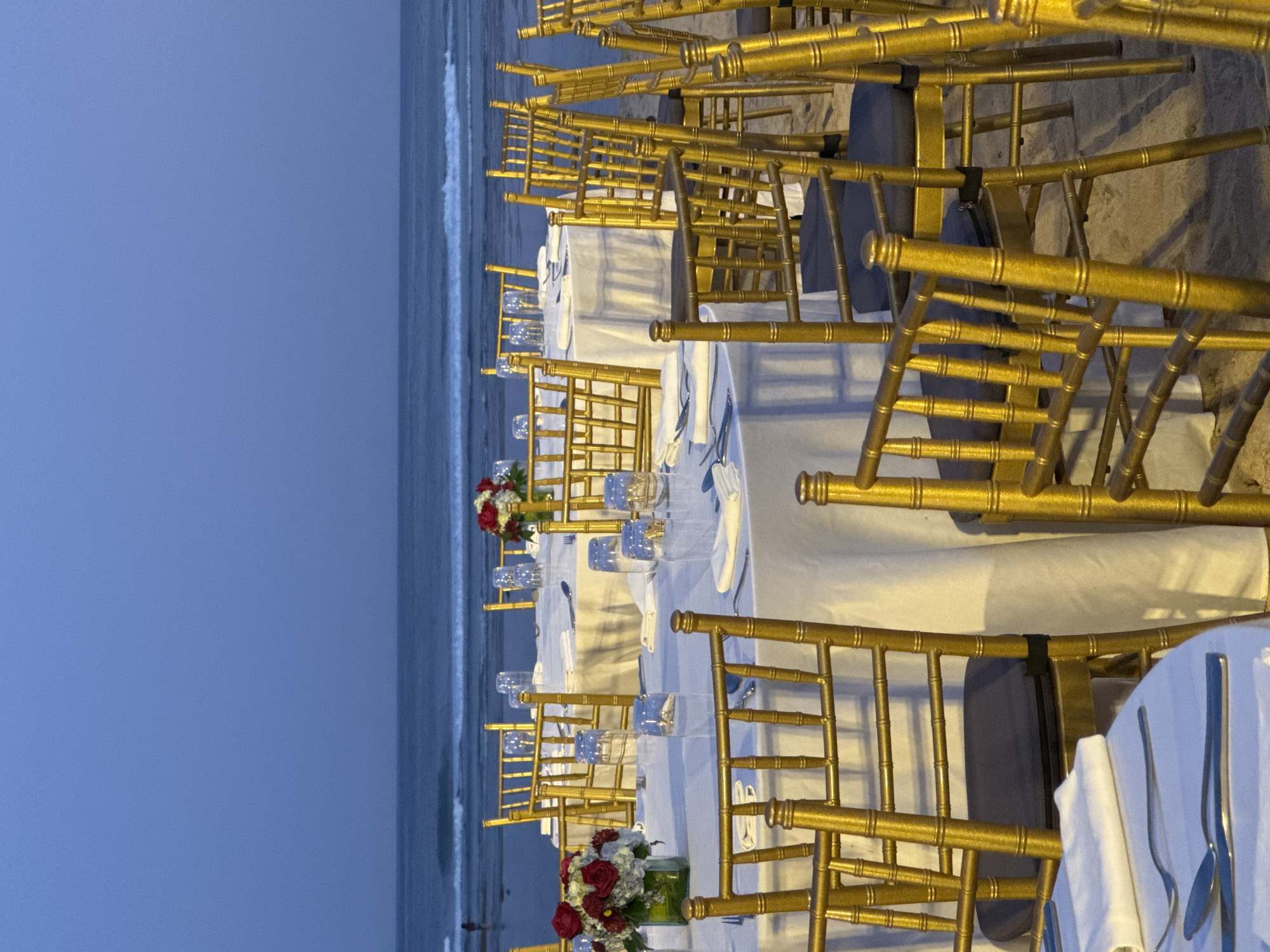 The image shows a row of tables set for a formal event on a beach in Kuta Selatan, Indonesia. The tables are covered with white linens and set with silverware, glassware, and centerpieces of red and white flowers. The chairs are gold and arranged around the tables. The beach is visible in the background, with the ocean and sky. The sky is a dark blue, suggesting it is evening or night.