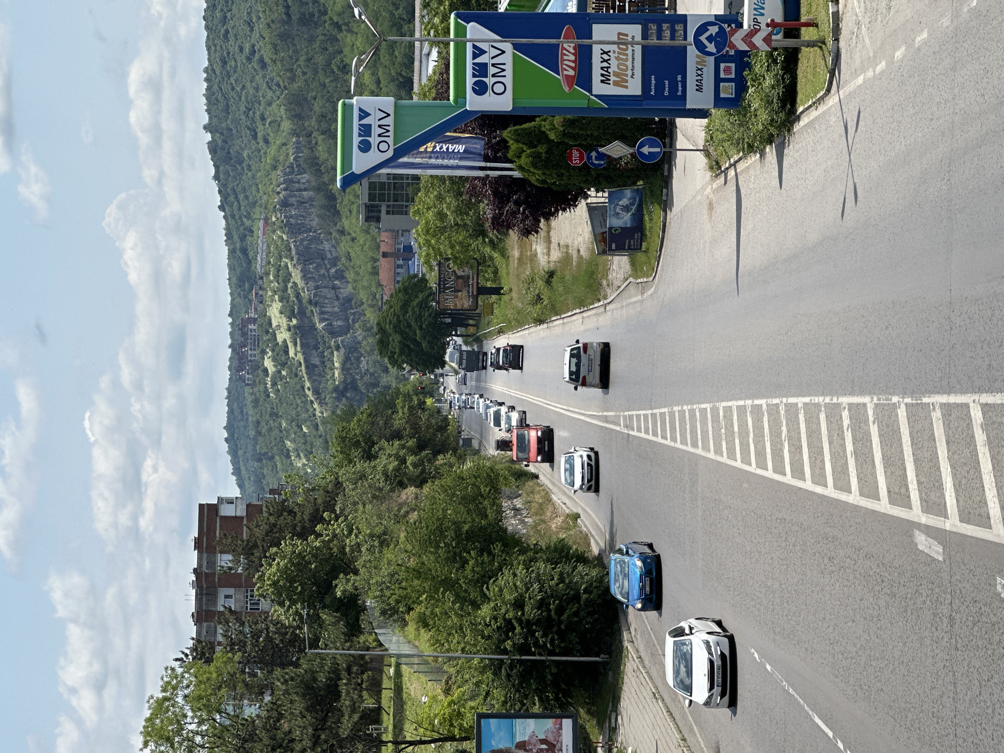 An overhead view captures a busy road curving slightly to the left, with traffic visible in both directions, extending into the distance. On the right side of the road, an OMV gas station is prominent, featuring large blue and green signs with its logo and fuel price displays. The station's signs advertise "VIVA" and "MAXX Motion Performance FIs," along with prices for "Autogas" (0.72), "Diesel" (1.64), and "Super 95" (1.56). A red "STOP" sign and a blue arrow sign are also visible near the gas station entrance.
To the left of the road, a dense, green mountain or hill covered in trees dominates the background. Rocky outcrops are visible on parts of the hillside, and a few multi-story buildings with brick facades are nestled among the greenery on the left side of the frame. The sky is partly cloudy with white cumulus clouds.
The scene depicts a clear, sunny daytime, likely late morning or early afternoon, given the shadows cast by poles and signs. Cars of various models and colors are present on the road; some appear to be moving, while others form a queue in the distance. A blue car and a white car are prominent in the foreground. There are billboards along the roadside, including one partially visible with the text "BIANCO." The setting suggests a main thoroughfare near a mountainous area, consistent with the provided location context of Veliko Tarnovo, Bulgaria.