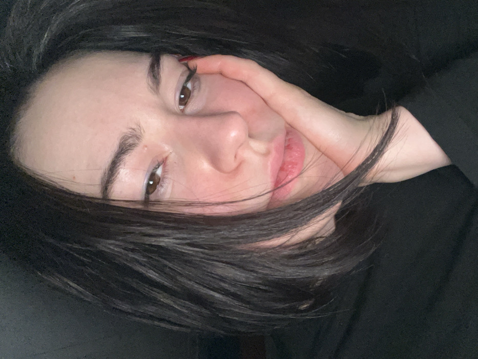 A woman with dark hair and light skin poses with her hand touching her face. She has brown eyes and full lips. Her hair is disheveled, partially obscuring her face. The image is taken at a close-up angle, with the woman's face angled towards the camera. A small glimpse of red nail polish is visible on one of her fingers. The background is dark, creating a high-contrast effect. The overall lighting is dim and moody.