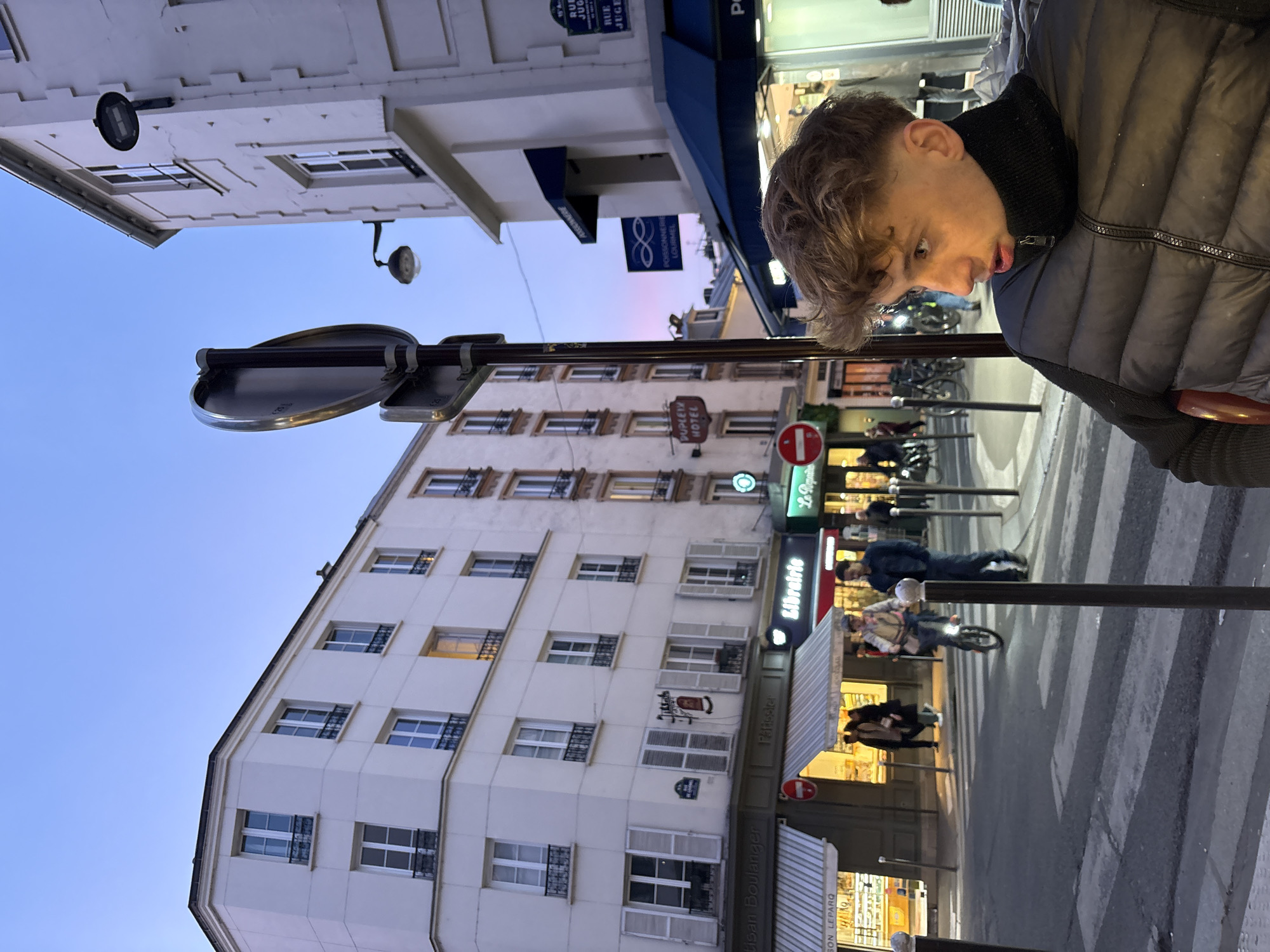 The scene depicts an urban street intersection in Paris, France, at dusk. The sky transitions from light blue overhead to a pink-orange gradient near the horizon.
In the immediate foreground, a young male, appearing to be in his late teens or early twenties, is seated, facing right and slightly forward. He has light brown, wavy hair and wears a black puffer vest over a black turtleneck. A dark vertical pole partially obscures the left side of his face.
The midground displays a pedestrian crossing with white stripes on dark asphalt. An individual on a unicycle, wearing a light-colored jacket and cap, is moving across the street from left to right, alongside another person walking. Multiple other pedestrians are visible on sidewalks and near storefronts.
Building facades, characteristic of Parisian architecture, are light-colored with multiple stories and numerous windows, some featuring white shutters and black iron balcony railings. Ground-level shopfronts are illuminated. Identifiable businesses include an "Artisan Boulanger / Patissier" with striped awnings on the left, a "Librairie" (bookstore) with a red illuminated sign, and a "Poissonnerie Gourmel" with a blue awning on the right. A red circular "no entry" sign is visible above the boulangerie awning.
Street signs in the upper right quadrant explicitly identify "RUE DU JOUR" and "RUE MONTMARTRE," locating the intersection in Paris. A sign for "DUPONT HOTEL" is visible on a building further down the street on the left. Streetlights are active, and several bicycles are parked along the street.