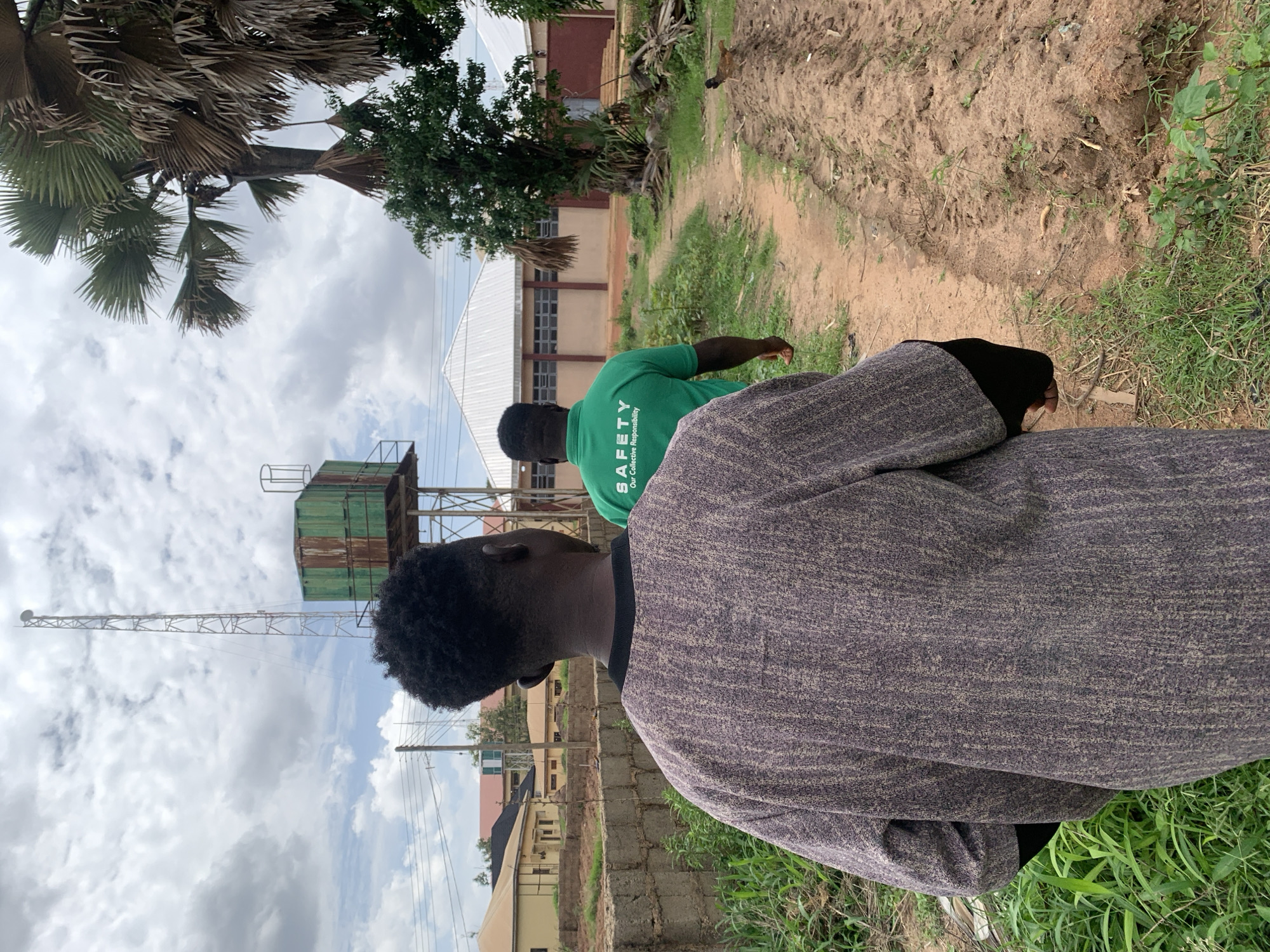 Two adult males are observed from behind, walking away from the viewer on an unpaved, reddish-brown dirt path in a semi-urban environment within the city of Jos, Nigeria.
The man in the immediate foreground, positioned slightly to the left of the frame's center, has short, dark, curly hair and is wearing a loose-fitting, long-sleeved garment with a mottled dark grey and brown textured pattern, featuring a black collar and black cuffs. His right arm is partially visible.
Ahead and slightly to the right of the foreground subject, a second man is visible, wearing a bright green short-sleeved t-shirt. The back of his shirt displays white text that reads "SAFETY Our Collective Responsibility."
The dirt path features varying textures and slight indentations, possibly from foot traffic or small vehicles. Green grass and low, leafy vegetation flank both sides of the path. To the right of the path, a raised earthen berm is visible, upon which a small brown and black chicken stands in the midground.
In the midground and background, various structures are present. On the left, several single-story buildings with light-colored walls and dark roofs are partially obscured by a low concrete block wall and some greenery. Overhead utility wires and poles are also visible on the left. In the central background, a prominent structure with a wide, light-colored gabled roof and brown walls with multiple windows is situated. To its left, a tall, slender metallic lattice tower, possibly a telecommunications mast, extends high into the cloudy sky, topped with an antenna. Adjacent to this tower, a large, rectangular, weathered green tank on a separate metal support structure is visible. Further to the right in the background, another building with a similar roofline to the central building is partially visible, alongside a substantial palm tree with broad, fanning fronds and other leafy trees. The sky is partially overcast with white and grey clouds, revealing some patches of blue.