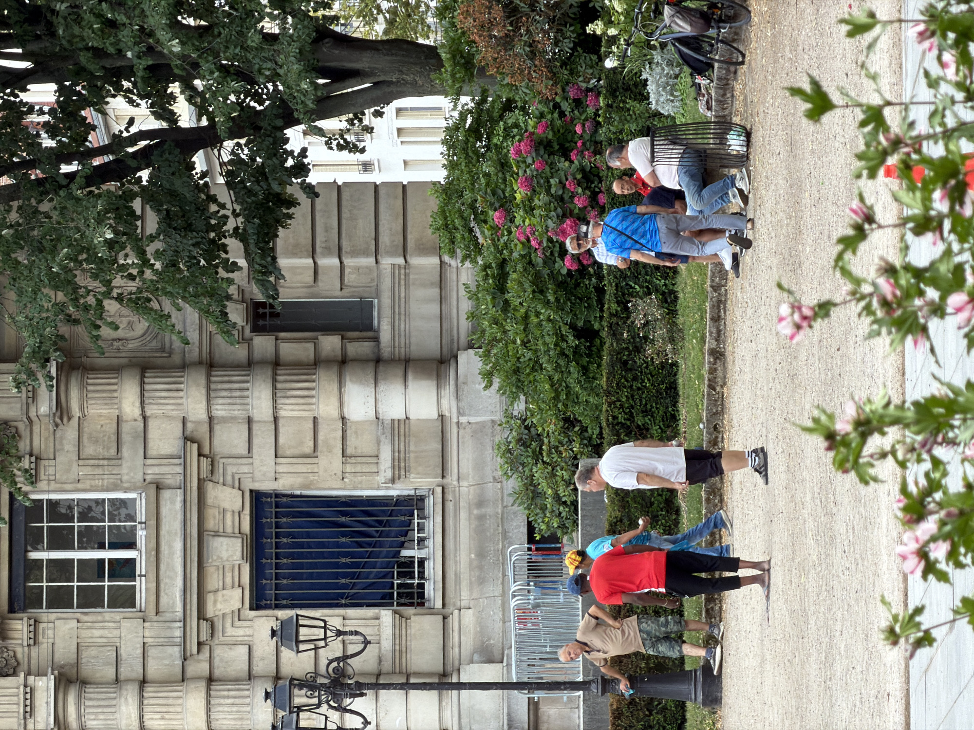 In Ivry-sur-Seine, France, on a sunny day, a group of men are playing a game of pétanque in a park with a gravel ground. The park is adjacent to a building with neoclassical architecture. Several men are gathered around a court, preparing to throw or watching the boules. One man in a red shirt and black pants is in the foreground, about to throw. To his left, another man in a tan shirt and camouflage shorts stands by a lamp post. To the right, a man in a white t-shirt and black shorts is observing the game. In the background, a dense green hedge and blooming pink flowers form a backdrop to the scene. Further back, a white building is visible through the trees. The overall atmosphere appears relaxed and social.
