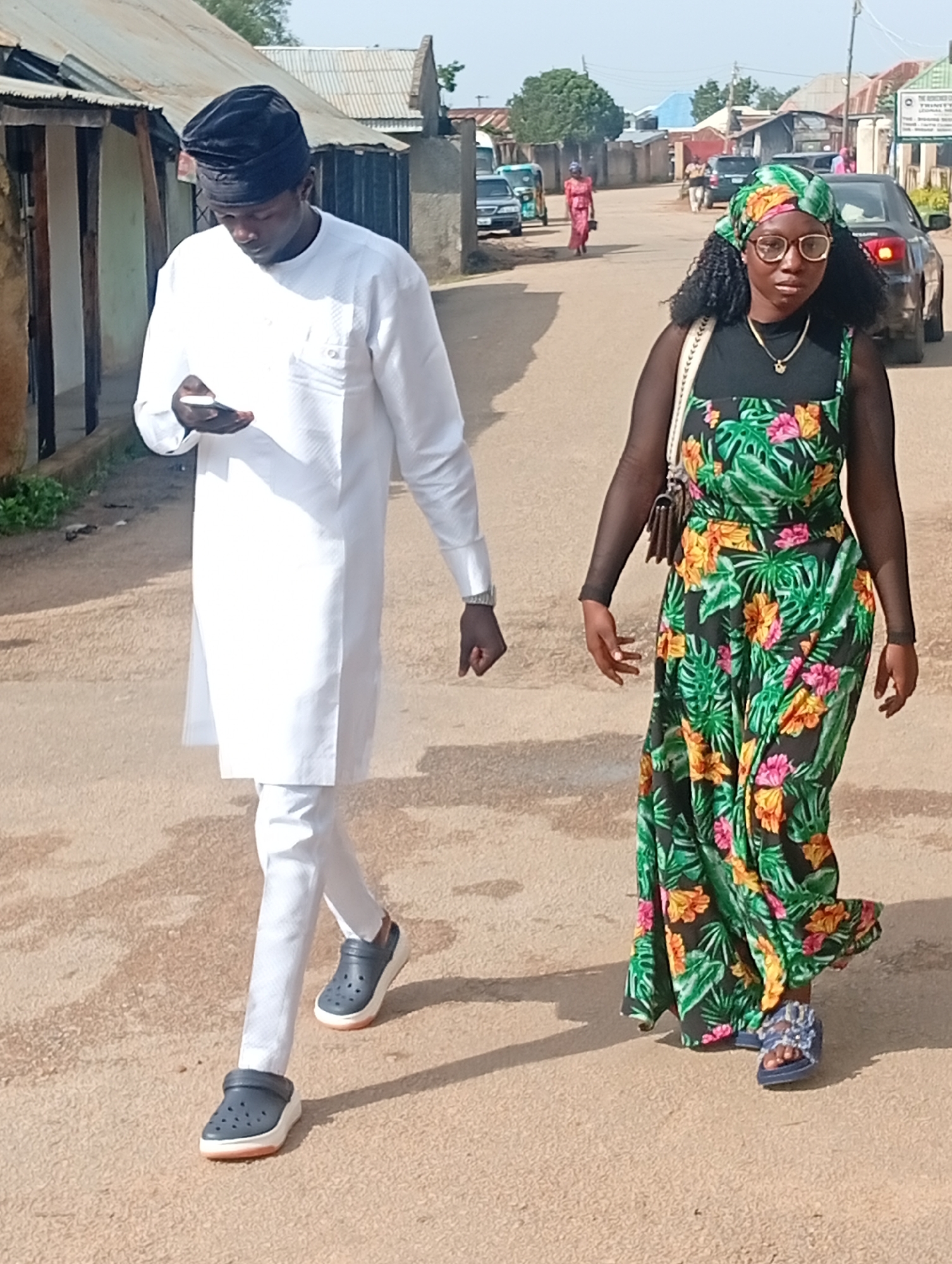 Two people, a man and a woman, are walking on a paved street in Jos, Nigeria. The man, on the left, wears a white traditional outfit and a black cap, and is looking at his phone. The woman, on the right, is wearing a floral-print dress and a green headscarf, and is carrying a handbag. In the background, there are buildings and vehicles.